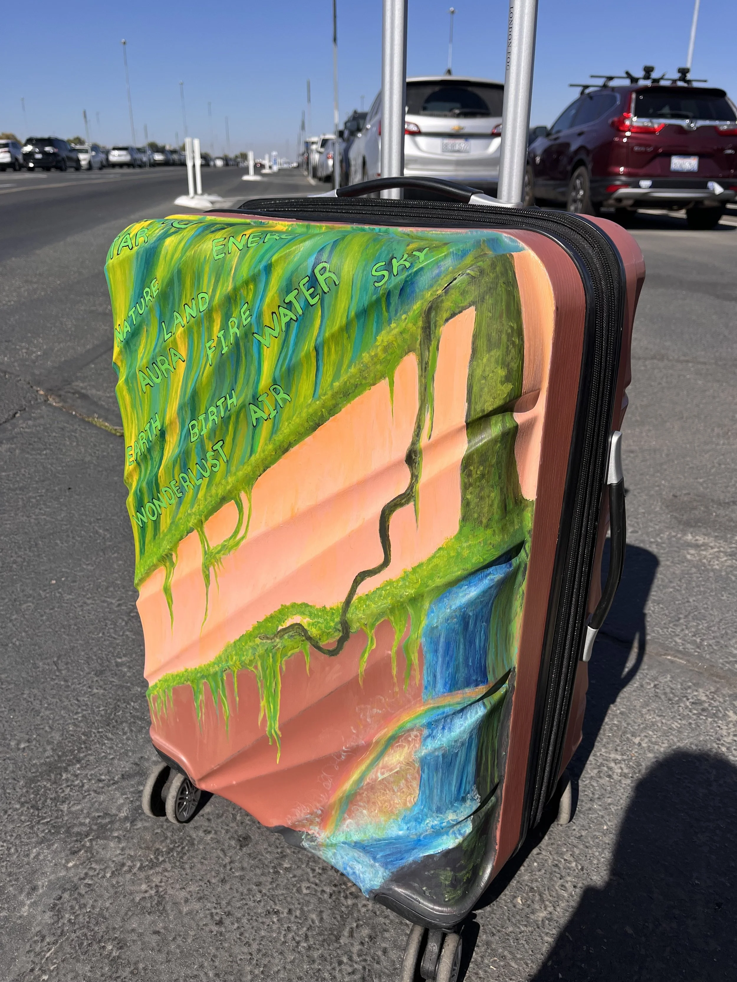Colorful suitcase with a painting of nature scene, featuring words like 'AIR,' 'WATER,' 'EARTH,' and 'SKY,' in a parking lot with parked cars and blue sky in the background.