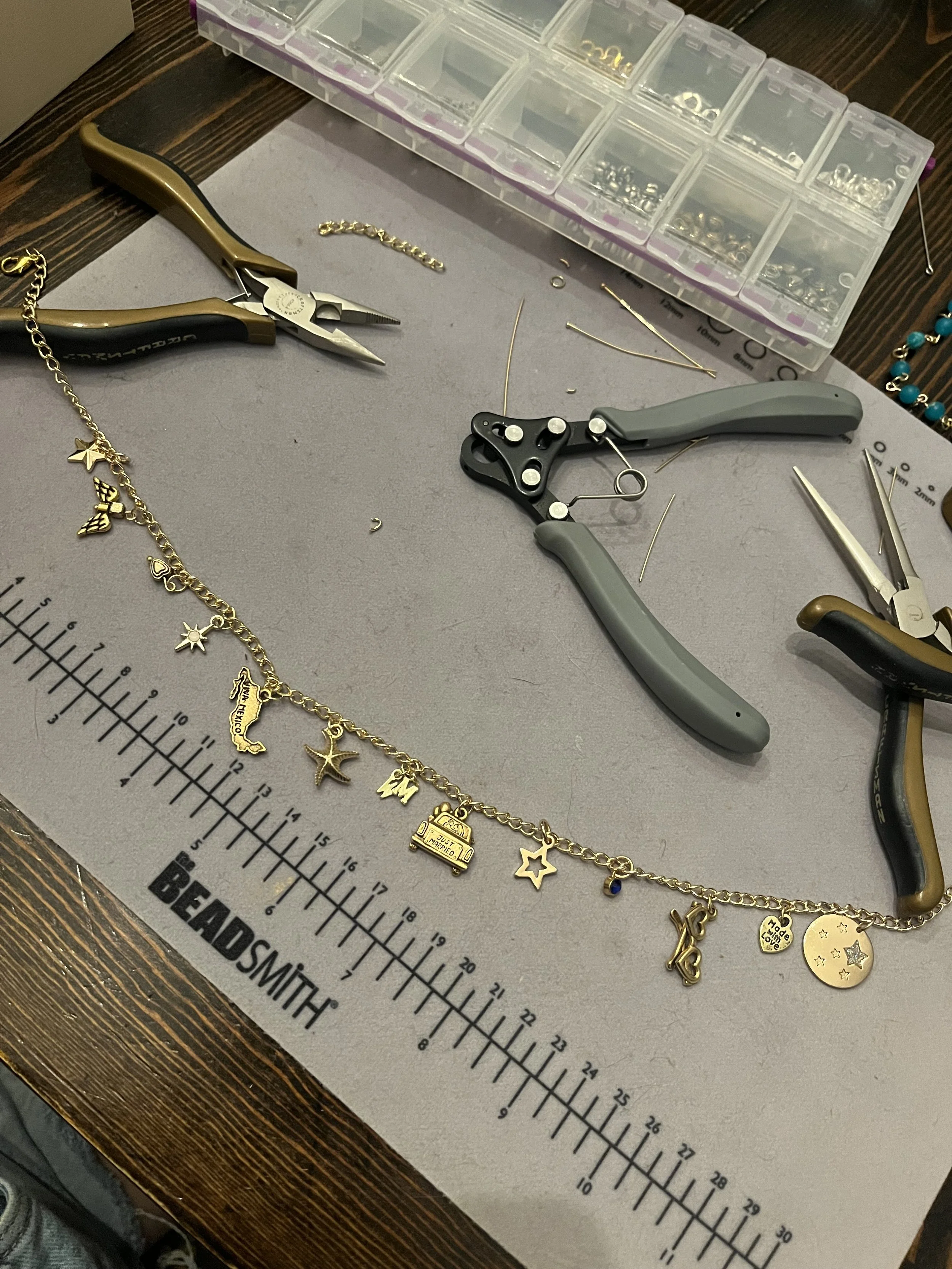 Jewelry making workspace with pliers, wire cutters, small parts, and a partially completed gold charm bracelet or necklace on a gray work mat with a Beadsmith measurement ruler.
