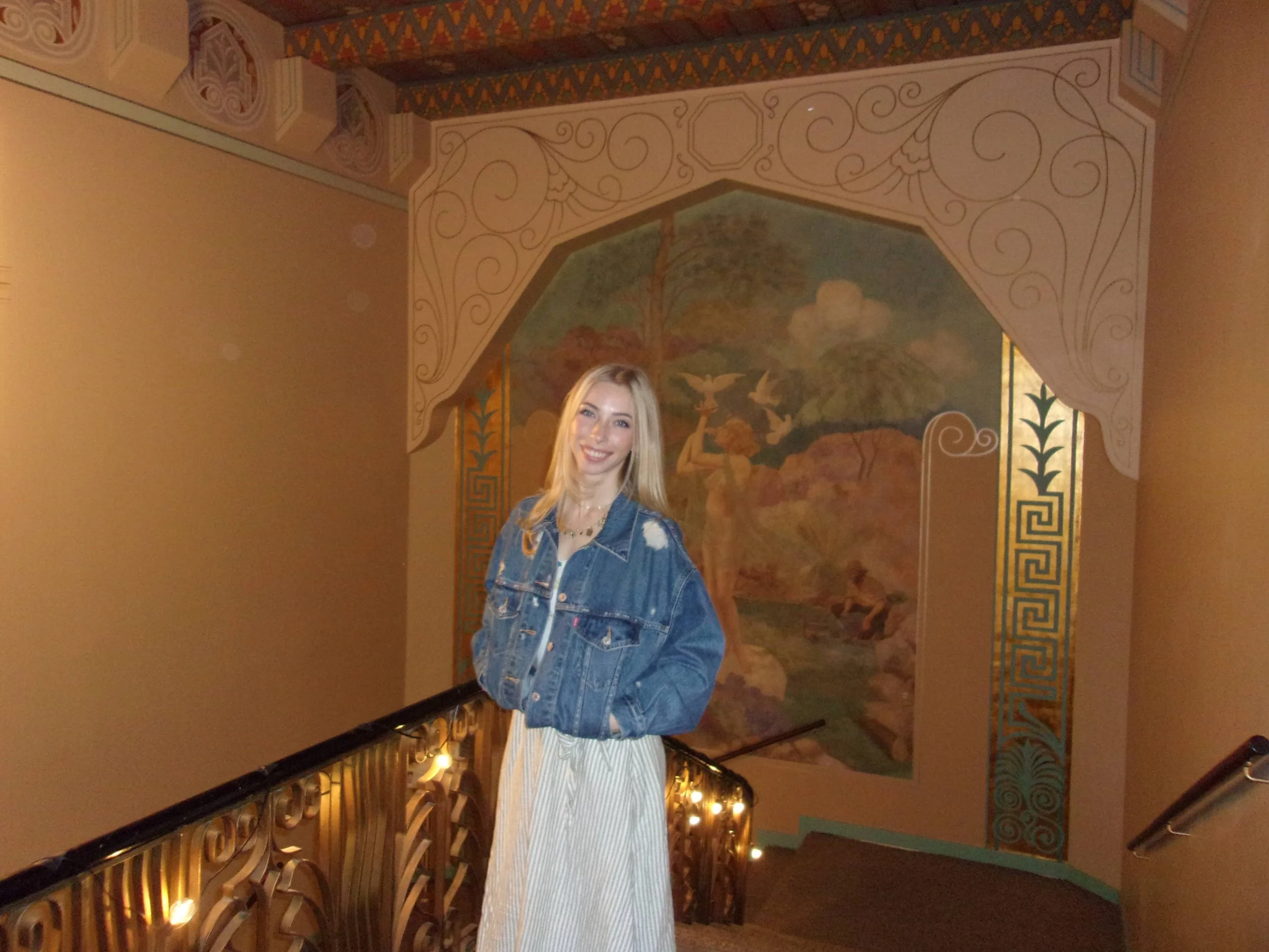 A smiling woman with long blonde hair wearing a denim jacket and a beige skirt stands on a staircase with ornate railing, in front of a mural of a woman with birds and trees, in a warmly decorated interior.