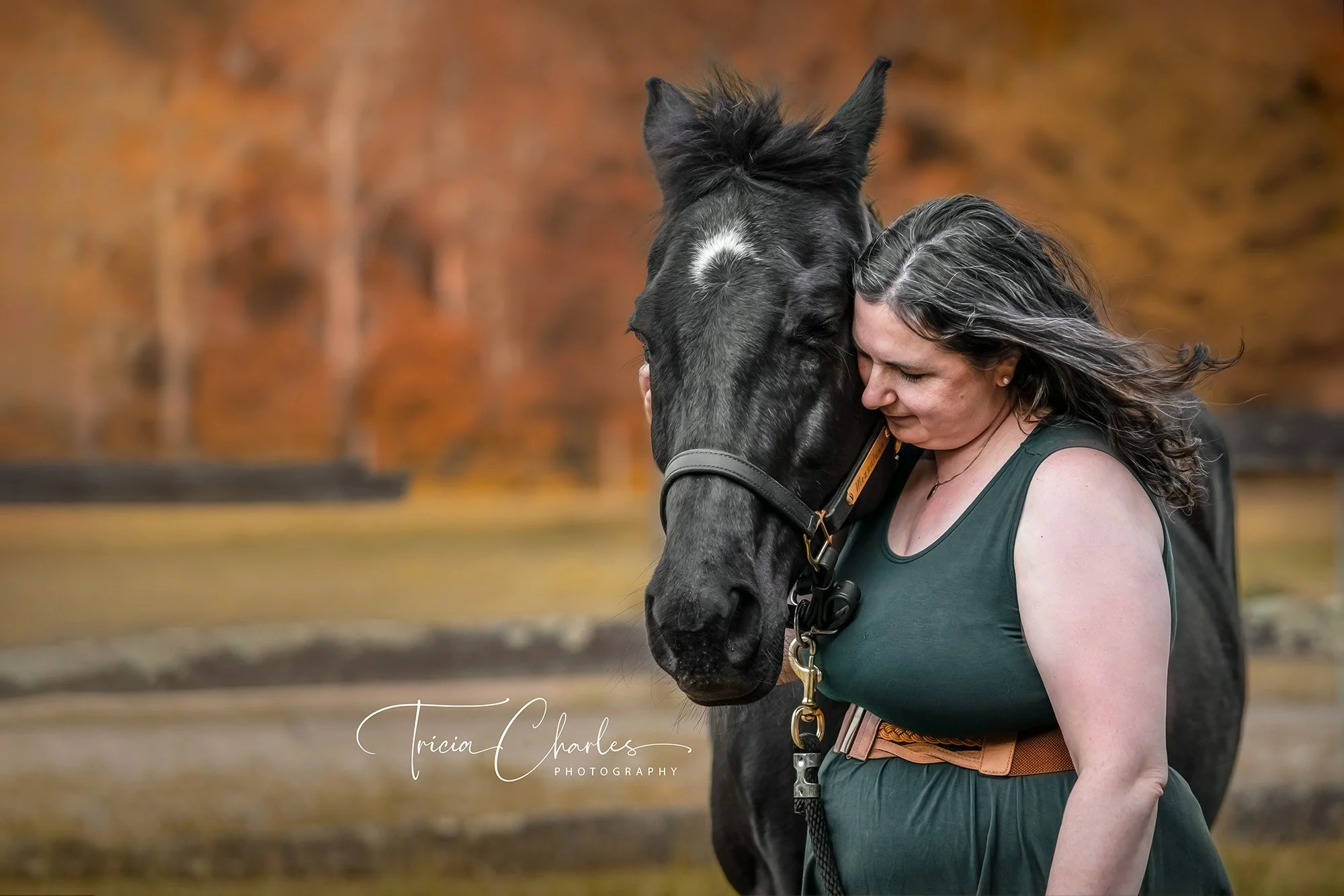 A woman with gray hair hugging a black horse outdoors with autumn trees in the background.
