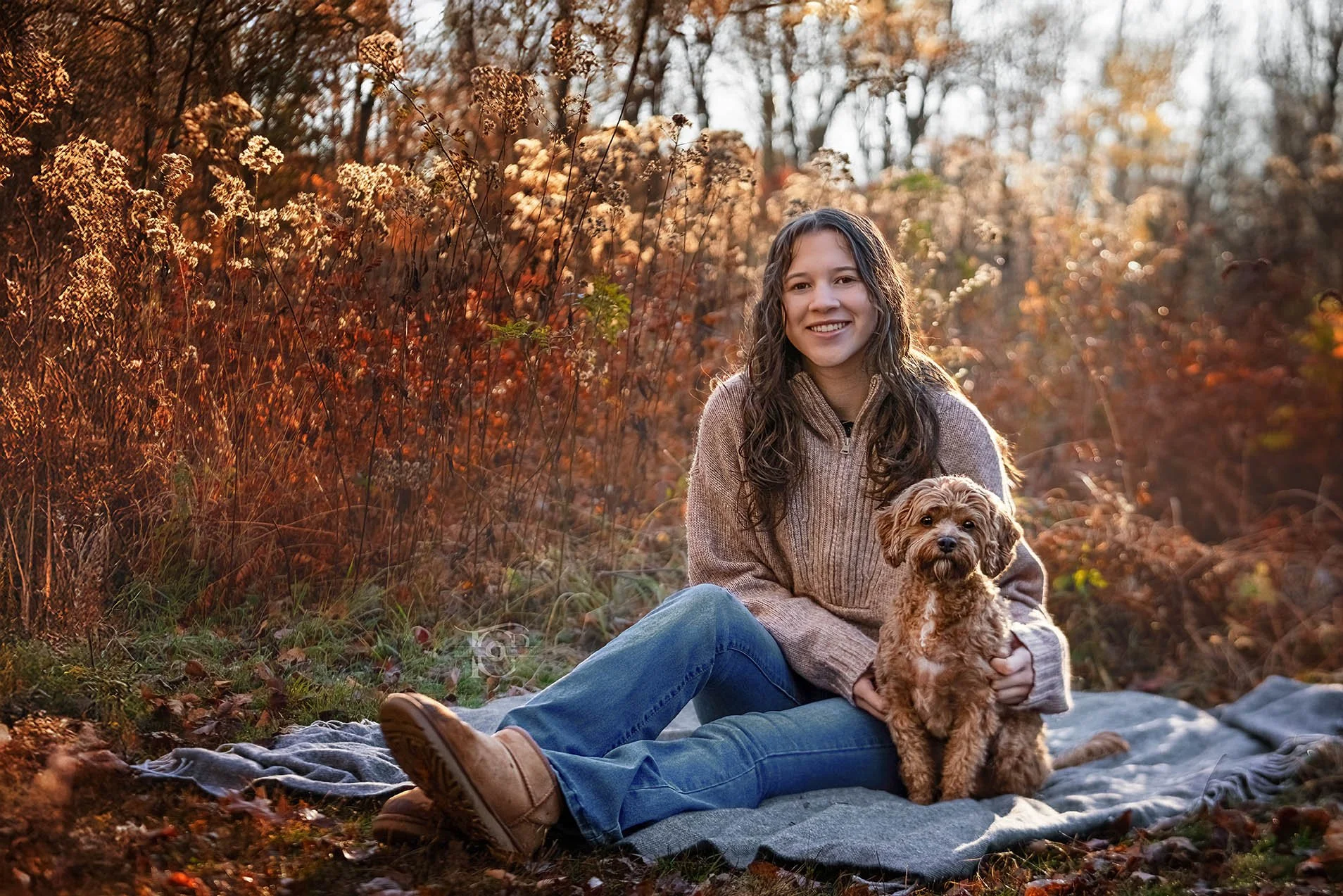 A woman with long hair smiling while sitting on a gray blanket with a small brown curly-haired dog, surrounded by autumn foliage in a forest during sunset.