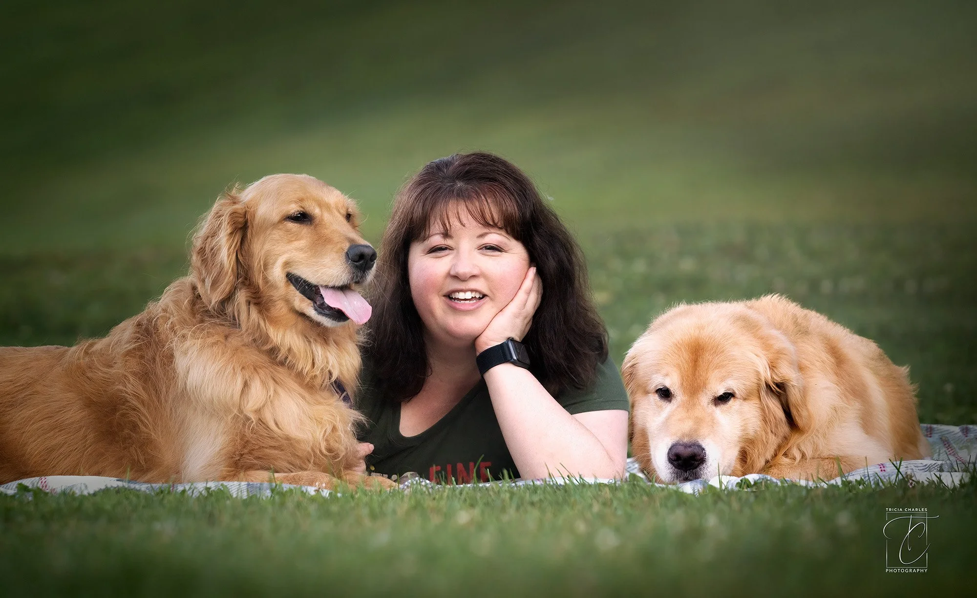 A woman with dark hair and a green t-shirt lying on a grassy field next to two golden retrievers, one panting and smiling, the other resting with eyes closed.