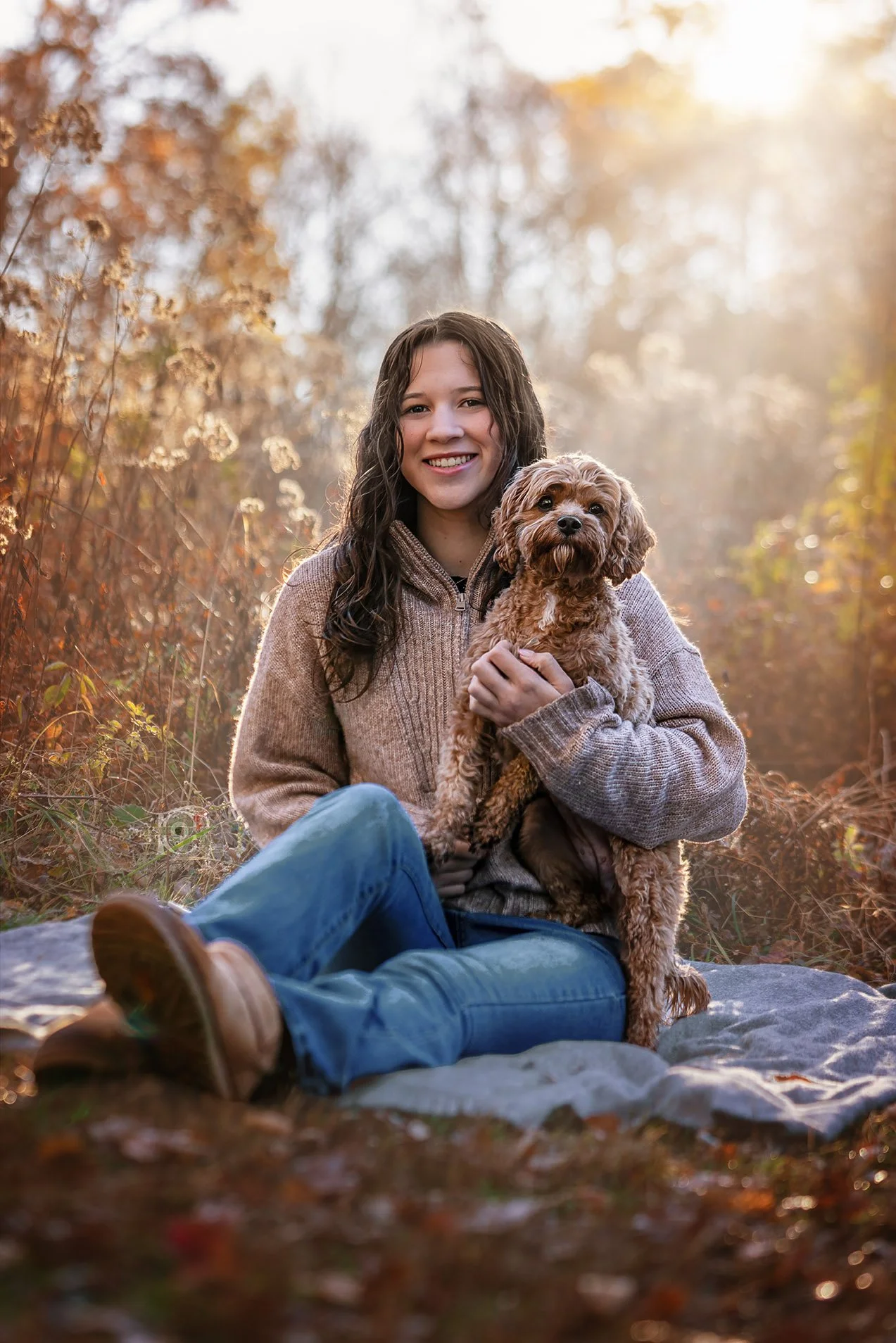 A smiling woman with long dark hair sitting outdoors in autumn, holding a small brown curly-haired dog. Sunlight filters through trees with orange and yellow leaves in the background. They are sitting on a blanket, with a grassy area in the foreground.