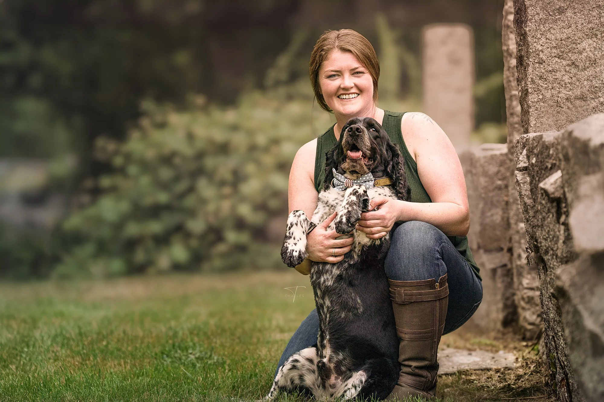 A woman with short brown hair and a green sleeveless top smiling while kneeling on the grass and holding a black and white puppy with long ears and a bow tie collar near a stone wall in a park.