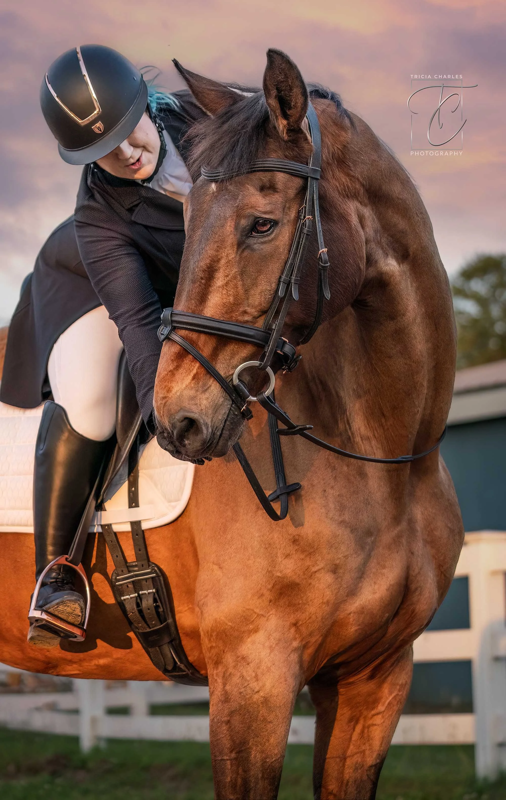 A person wearing a helmet and riding breeches on a brown horse with a white saddle pad at an outdoor riding arena during sunset.