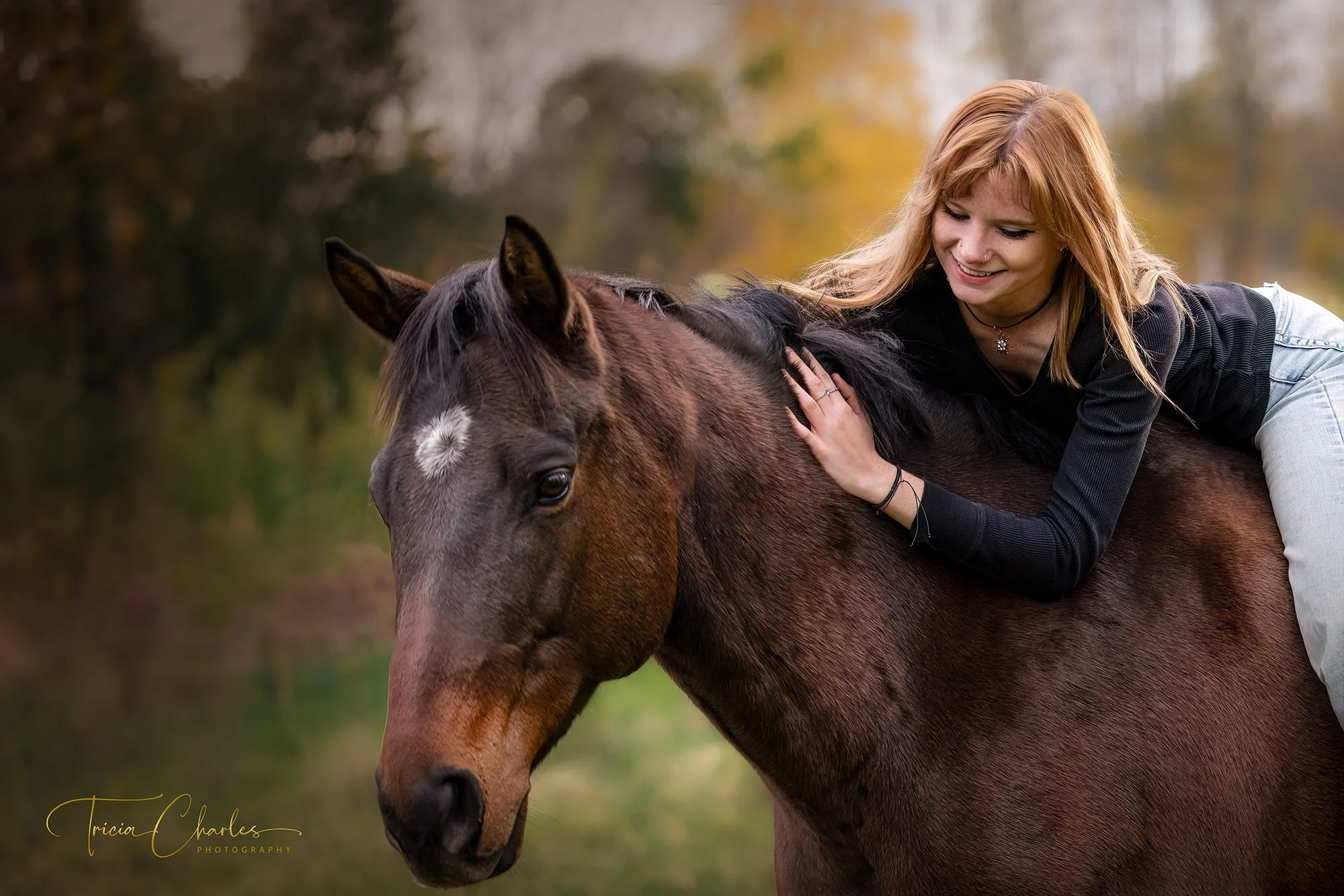 A woman with red hair, wearing a black sweater and multiple rings, is lying on a brown horse, petting its neck and smiling. The background features blurred autumn trees and foliage.