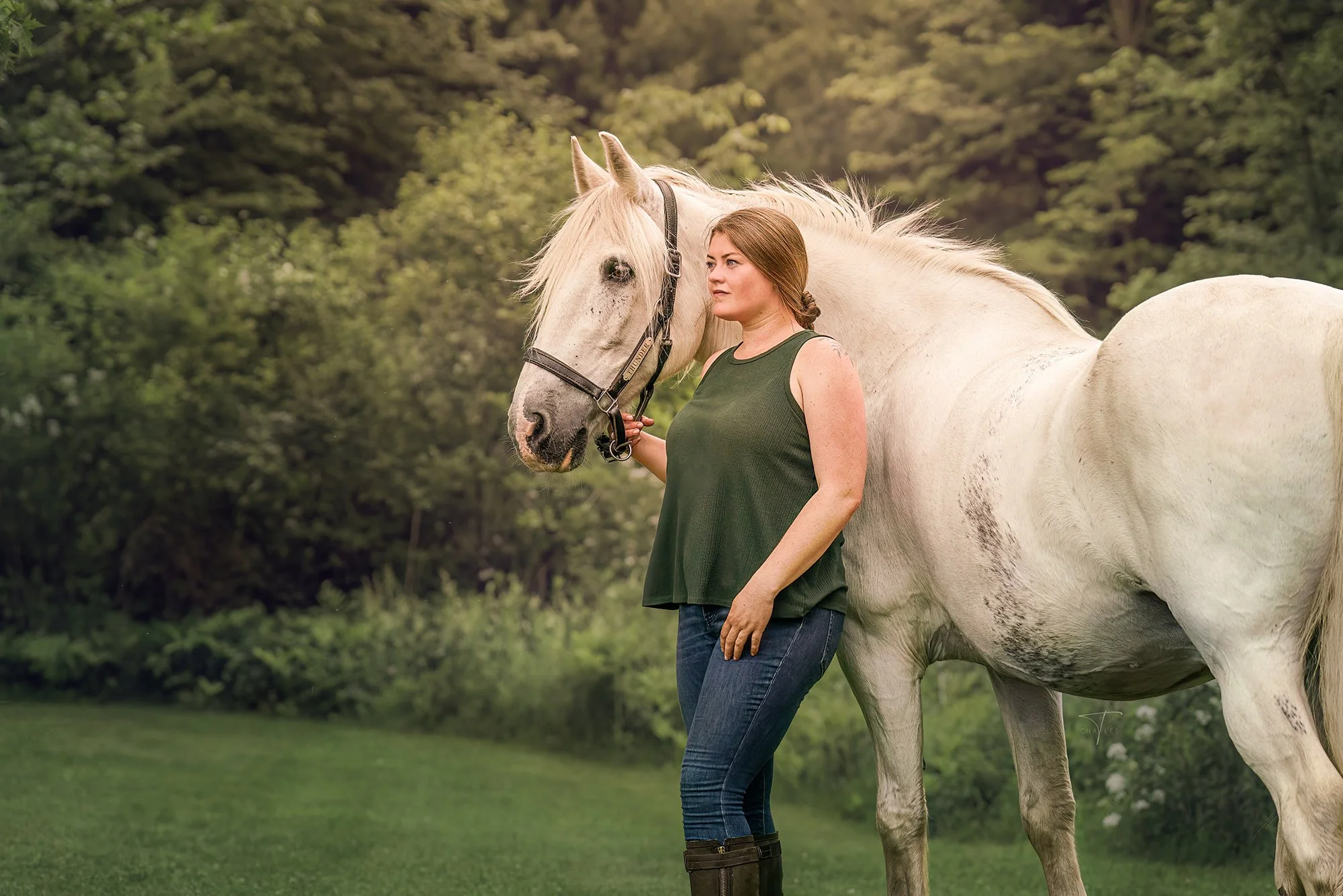 A woman with long hair in a bun standing next to a white horse in a green outdoor setting with trees in the background.