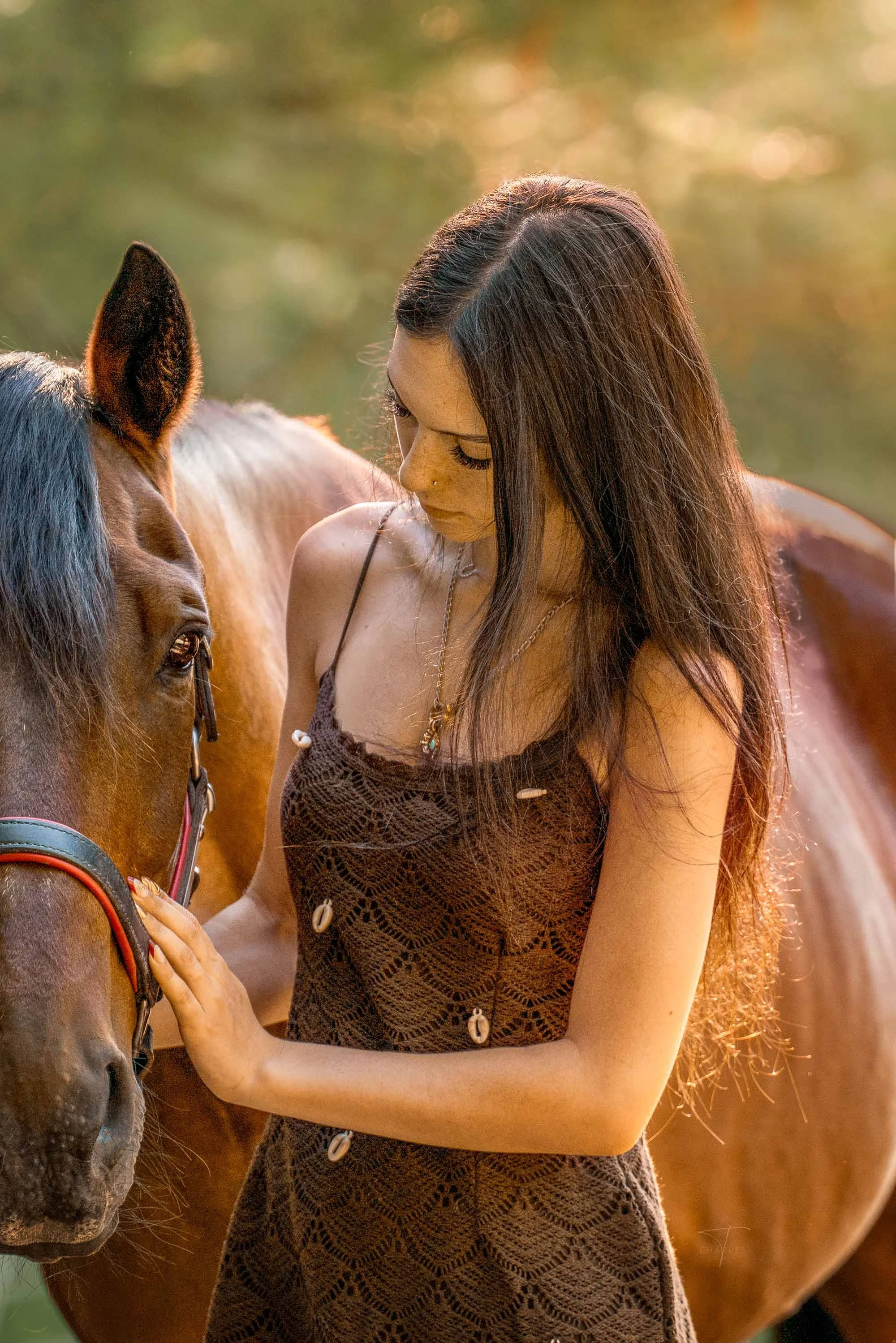 A young woman with shoulder-length blonde hair, wearing a black shirt with thin straps and a necklace, stands beside a dark brown horse with a black mane, in an outdoor setting with trees and a cloudy sky in the background.
