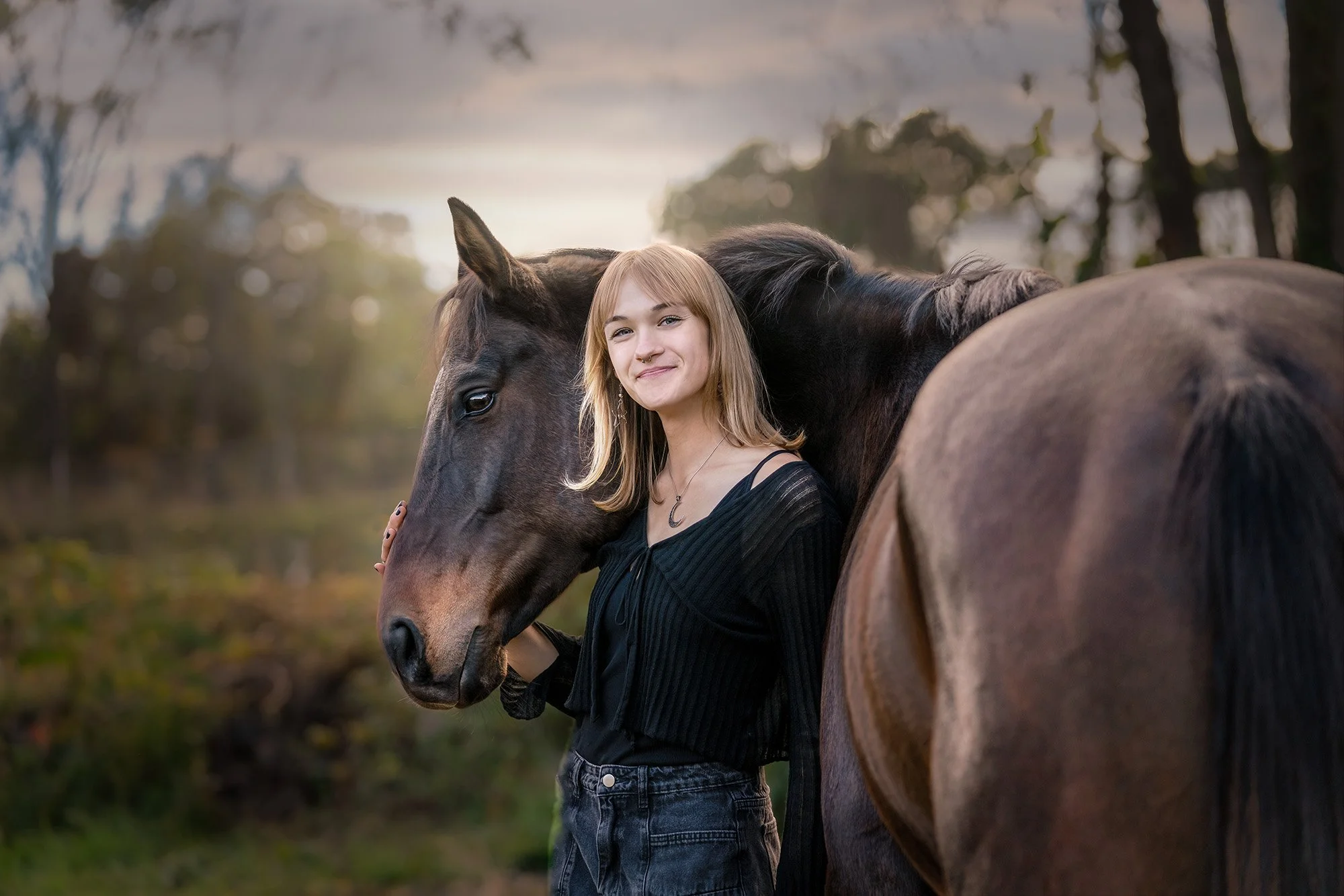 A young woman with shoulder-length blonde hair, wearing a black shirt with thin straps and a necklace, stands beside a dark brown horse with a black mane, in an outdoor setting with trees and a cloudy sky in the background.