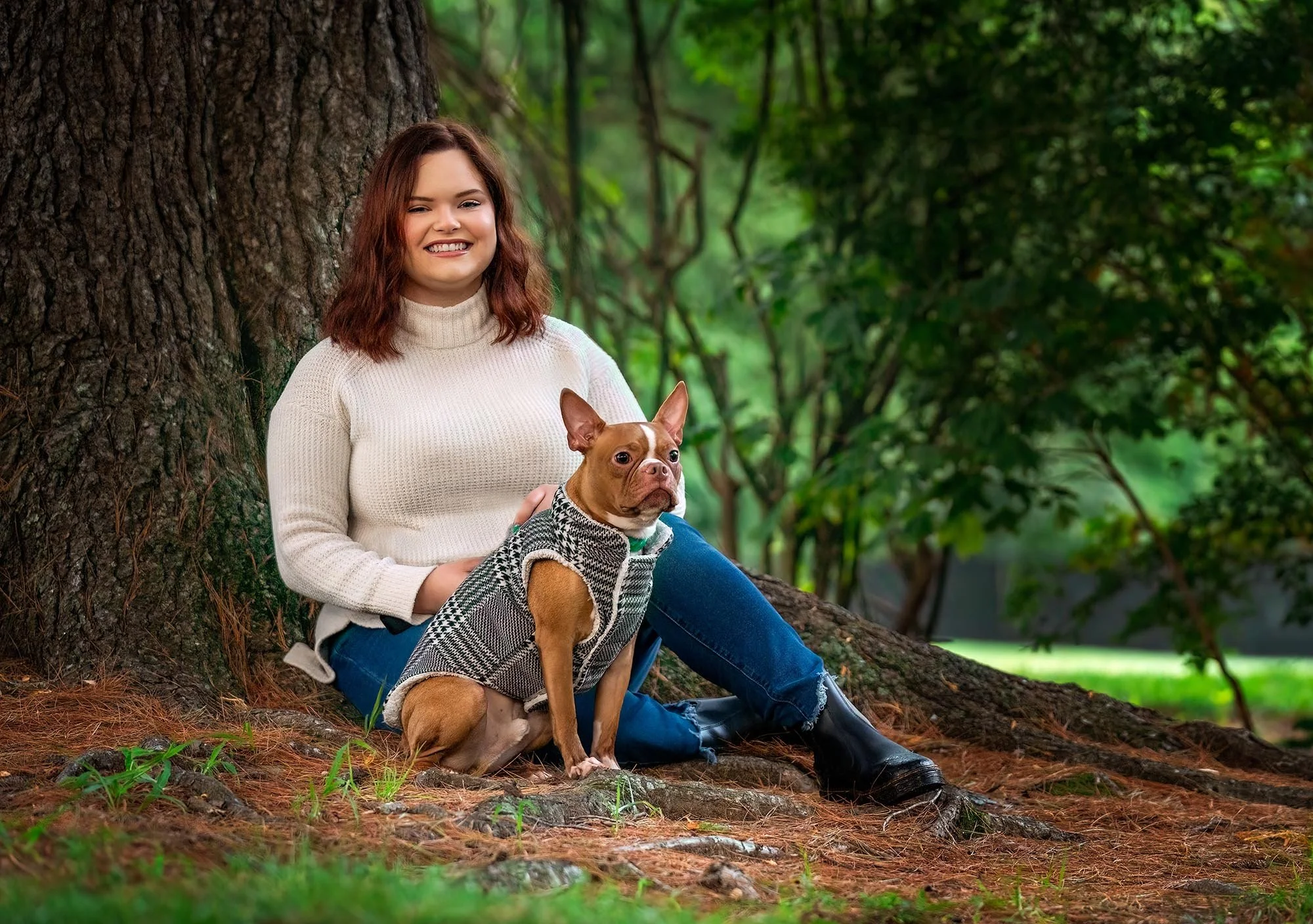 A young woman with wavy brown hair sitting on the ground next to a large tree, smiling, holding a small brown and white dog wearing a plaid coat, in a lush green park.