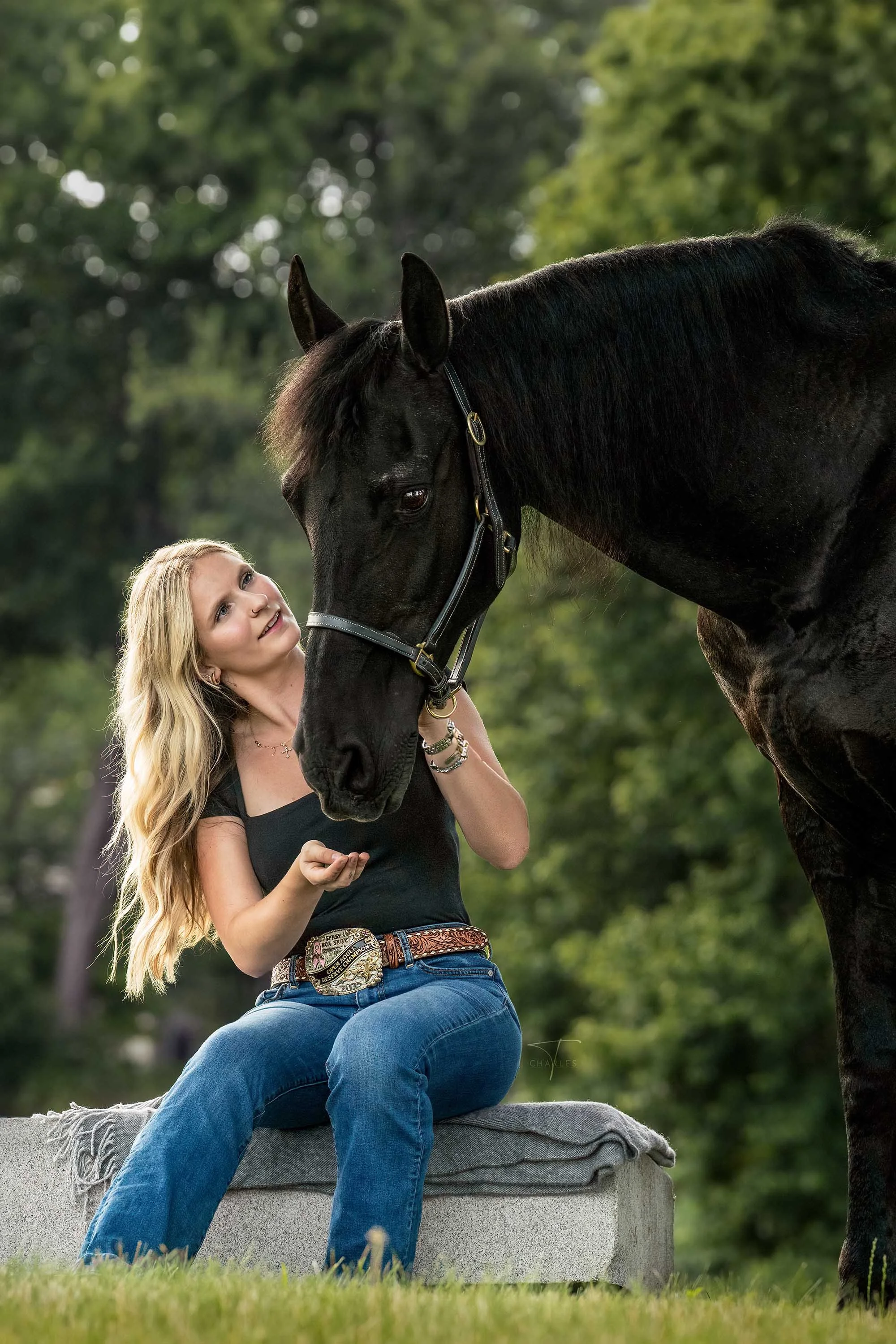 A woman with long curly hair kneeling in a grassy field, touching the nose of a white horse with a dark mane during sunset.