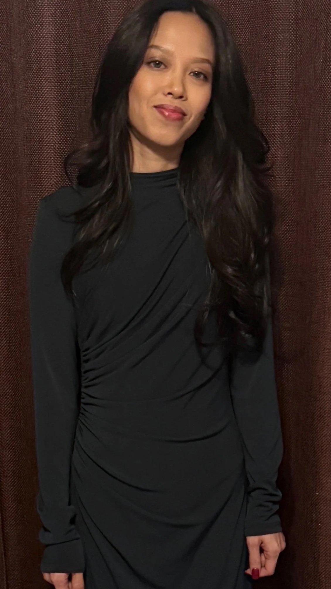 A woman with long dark hair wearing a black dress and red nail polish stands in front of a brown textured background.