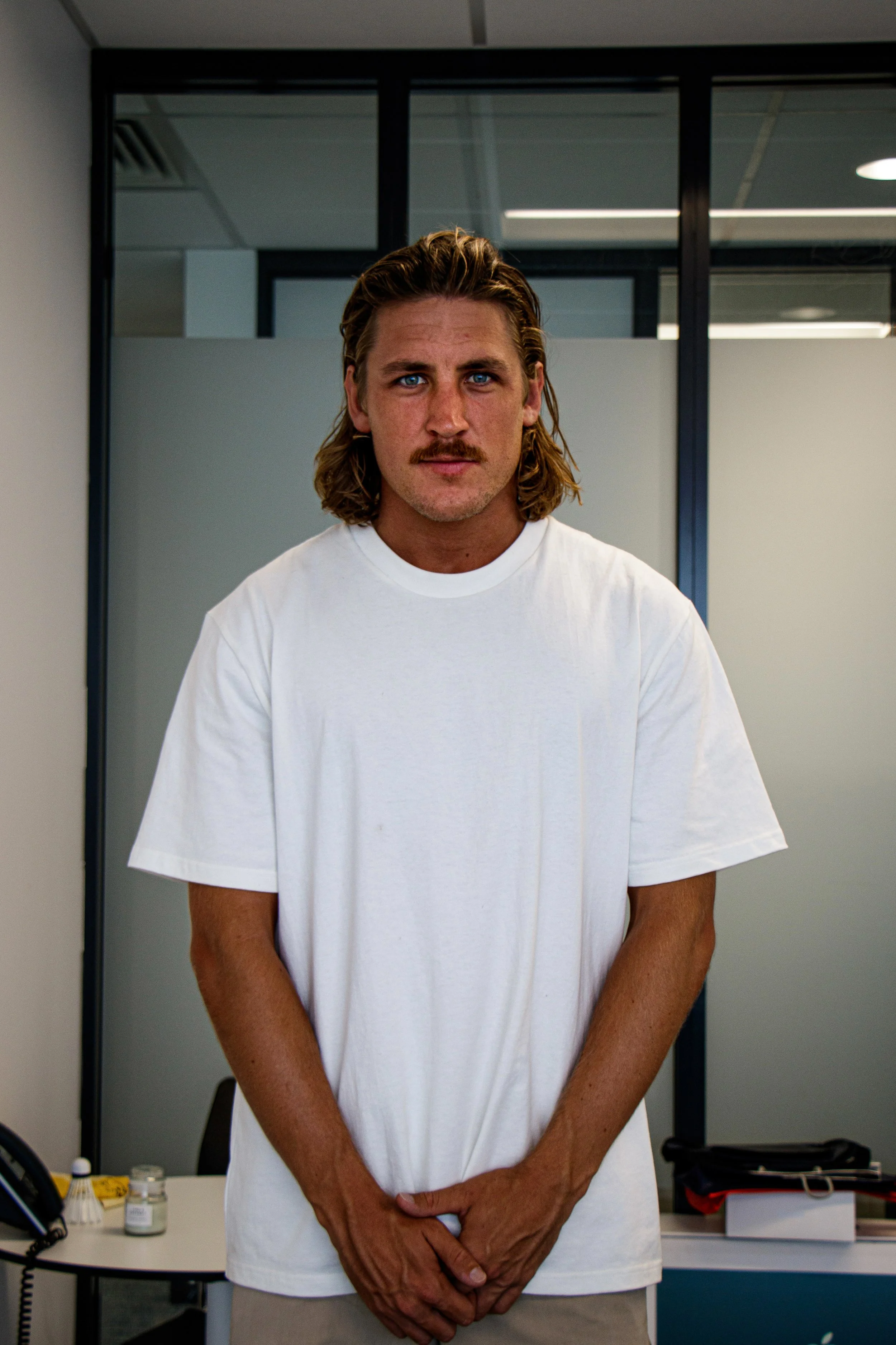 A man with shoulder-length wavy blond hair, blue eyes, and a mustache wearing a plain white T-shirt, standing in an office environment.