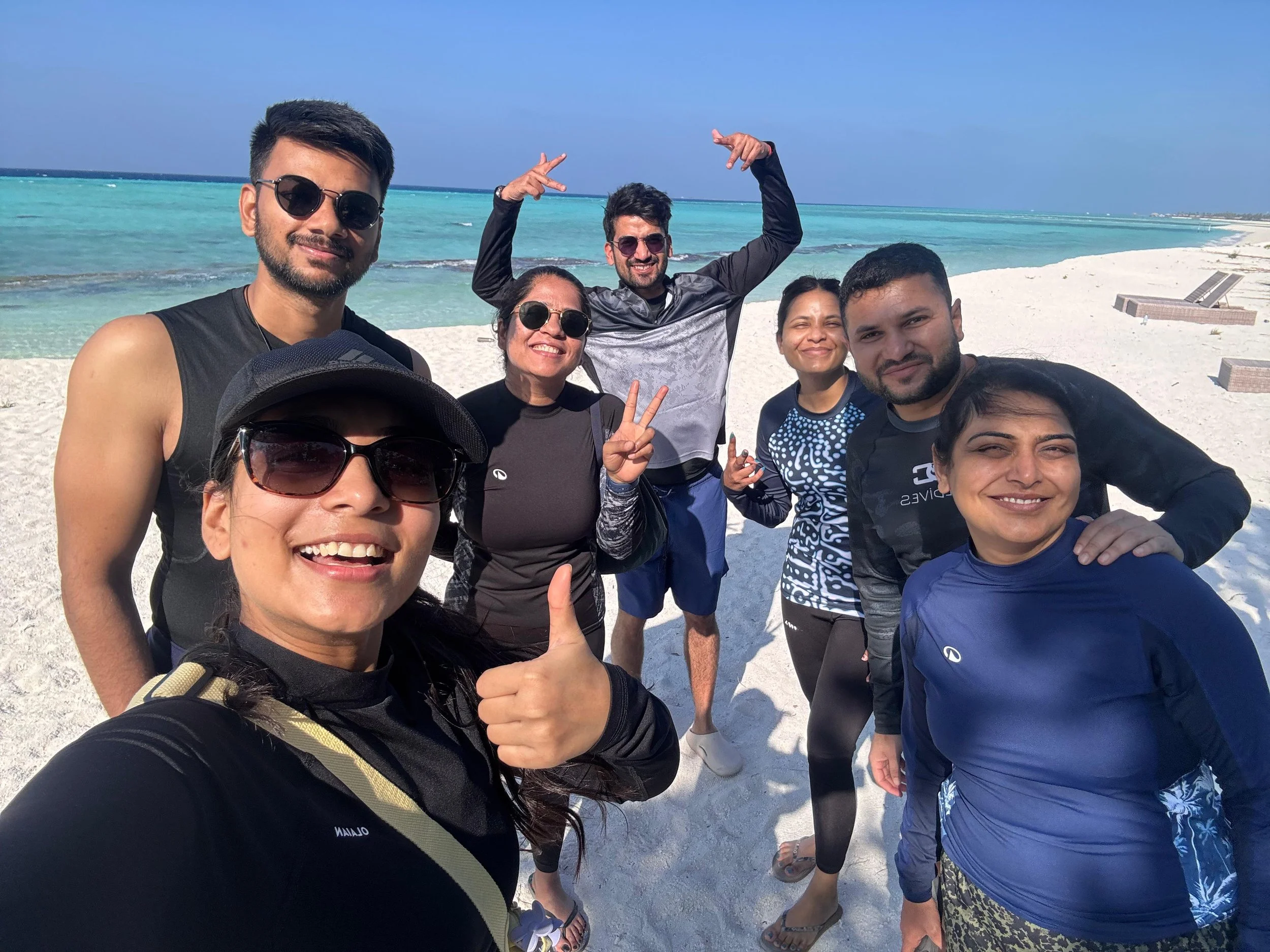 A group of eight friends smiling and taking a selfie on a white sandy beach with the turquoise ocean and blue sky in the background.