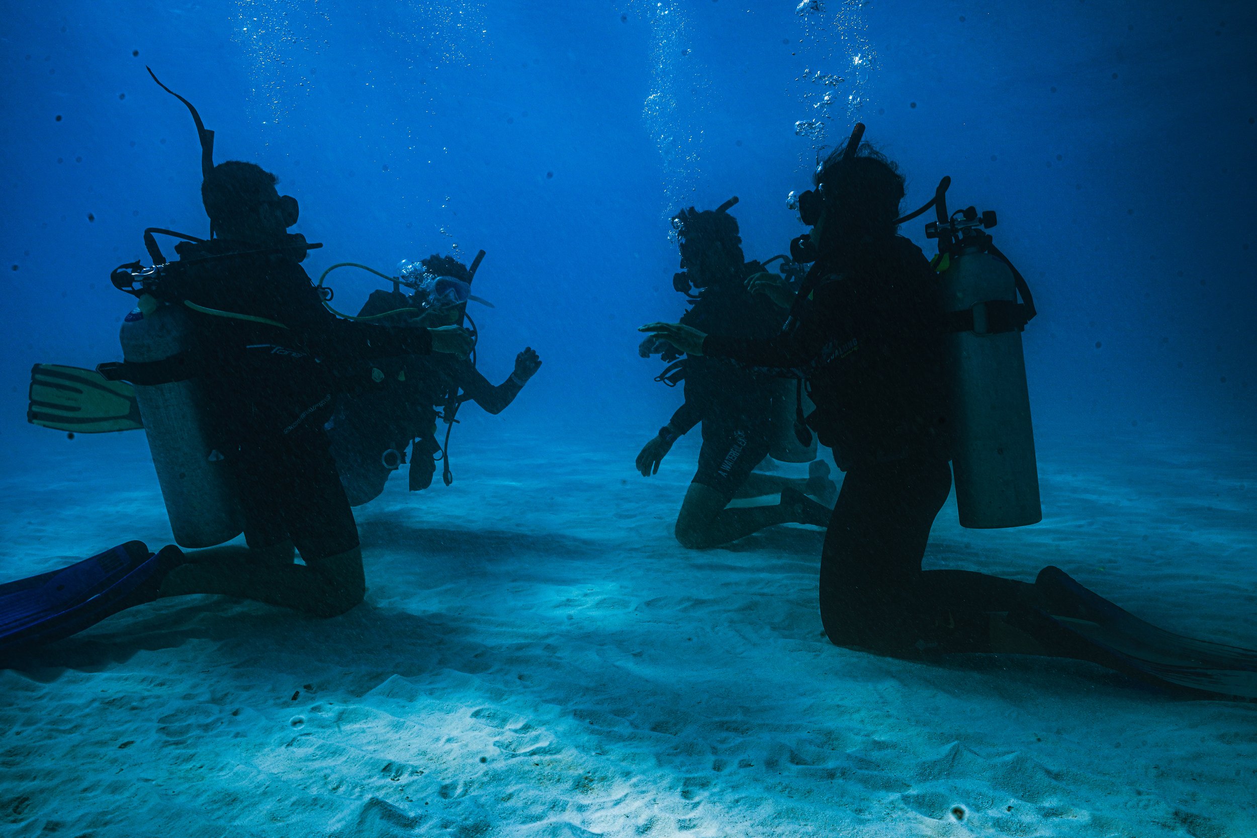 Four scuba divers kneeling underwater, wearing wetsuits and scuba gear, engaging in a training or rescue exercise.
