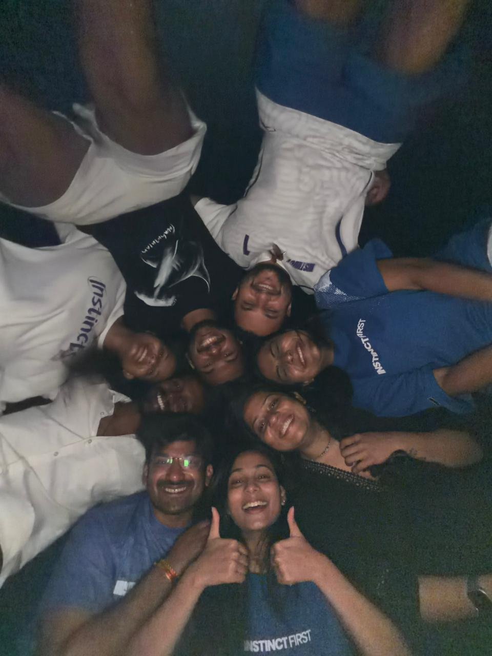 Group of ten smiling people lying in a circle with heads touching, taking a selfie and making thumbs-up gestures, indoors.