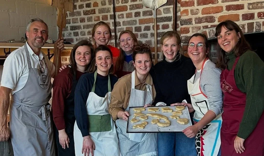 Groep van acht vriendinnen die trots lachen met het deeg dat ze maakten tijdens een teambuilding activiteit bij Bakker Baert vooraleer het de oven in gaat..