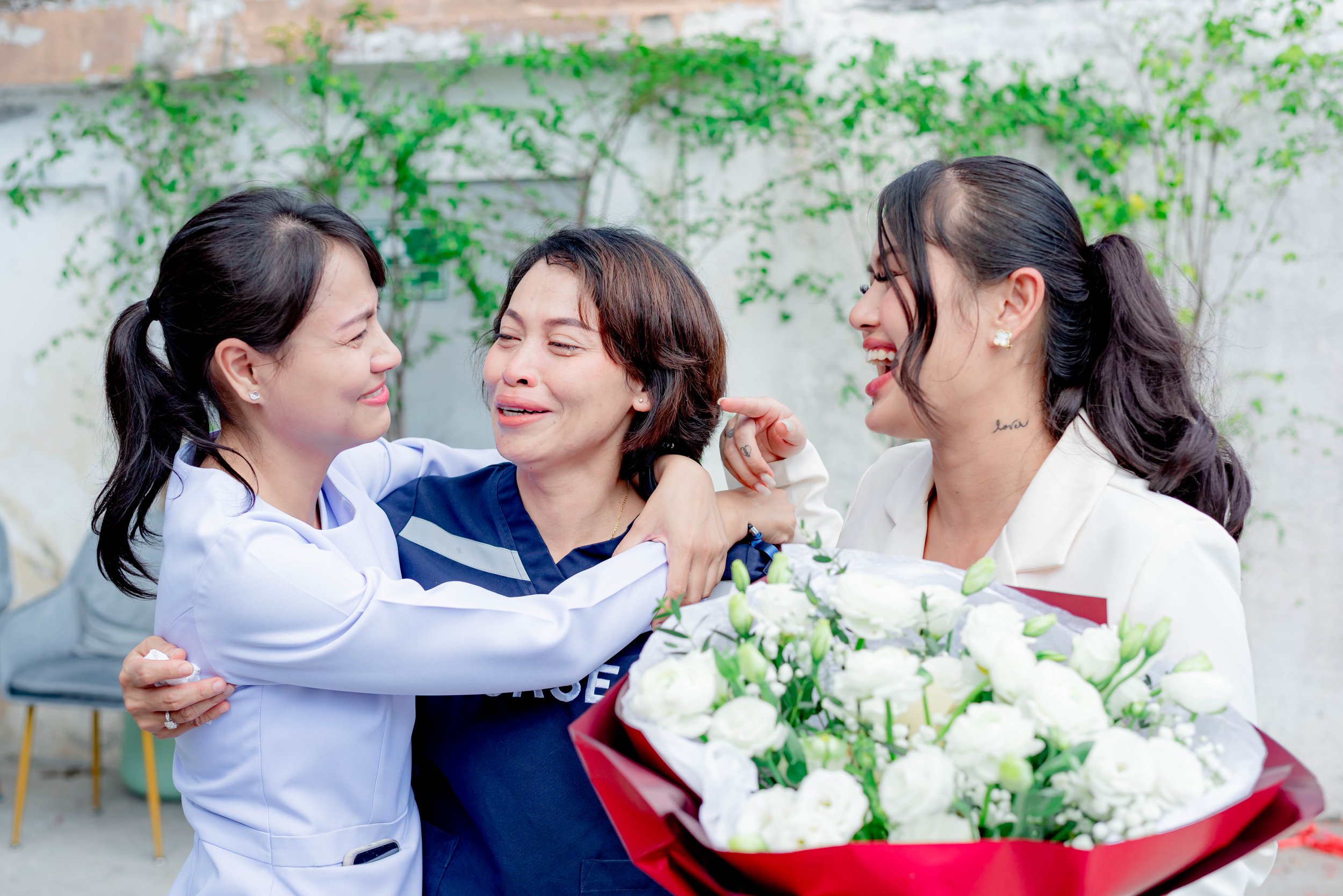 Three women smiling and embracing; one holds a bouquet of white flowers.