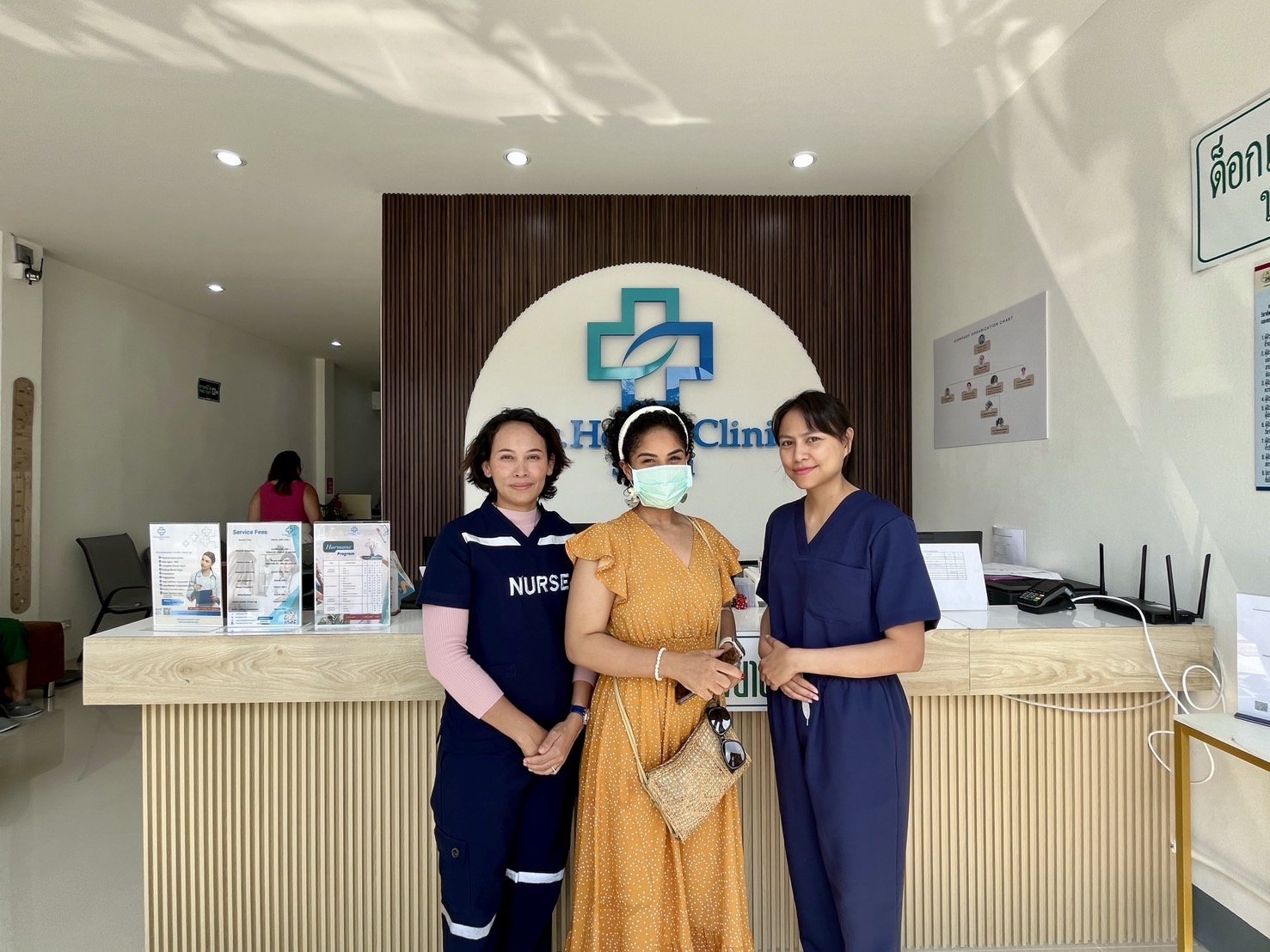 Three women standing in front of a clinic reception desk, with a woman in a nurse uniform and a woman in a mask. The clinic logo is visible on the wall.