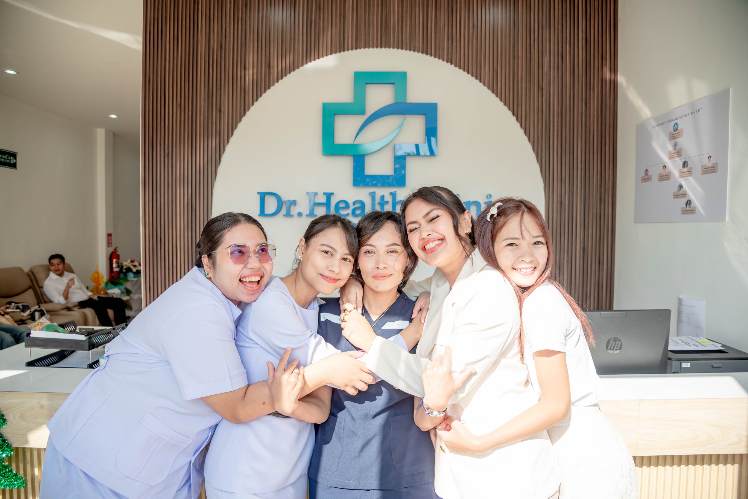 Group of five smiling women hugging in a healthcare clinic reception area, with a logo and sign reading "Dr. Health Clinic" in the background.