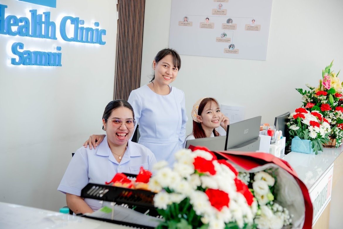 Reception area of a health clinic with three smiling staff members, a computer monitor, and flower arrangements.