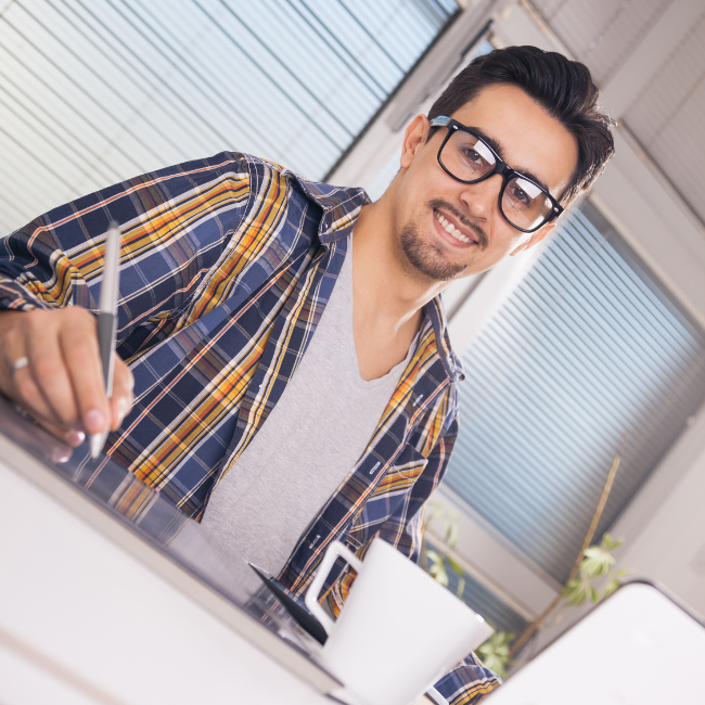Smiling man wearing glasses and a plaid shirt sitting at a desk in an office, holding a pen and working on a notebook with a coffee mug nearby.