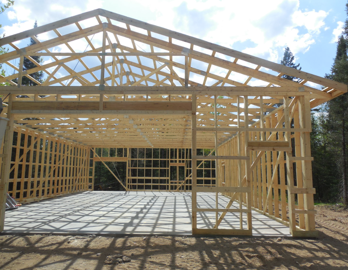 Wood frame structure of a house under construction with sunlight casting shadows on the concrete foundation, surrounded by trees and blue sky with clouds.
