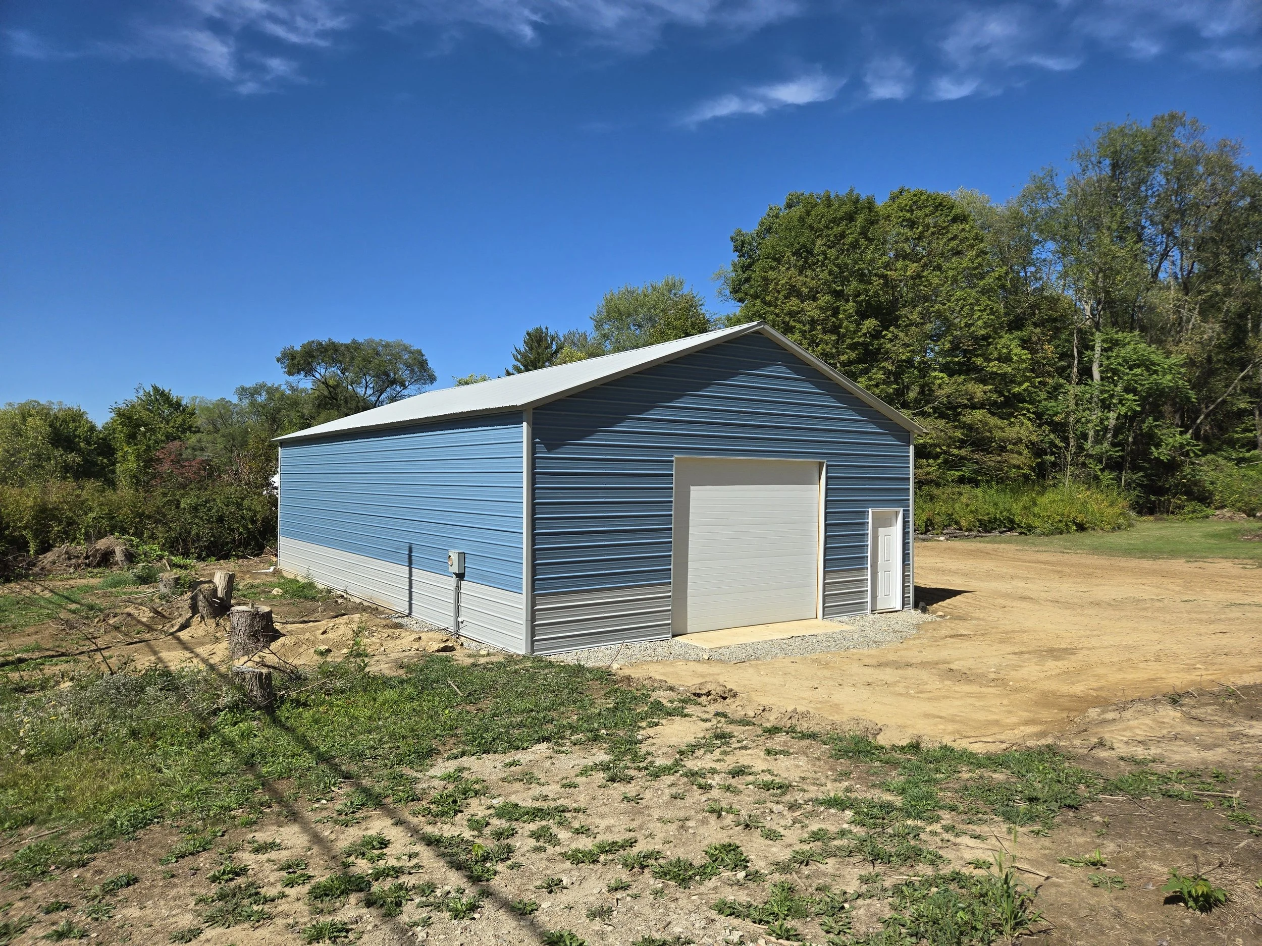 A blue metal shed with a white garage door and side door, situated on a dirt lot with a background of trees under a clear blue sky.