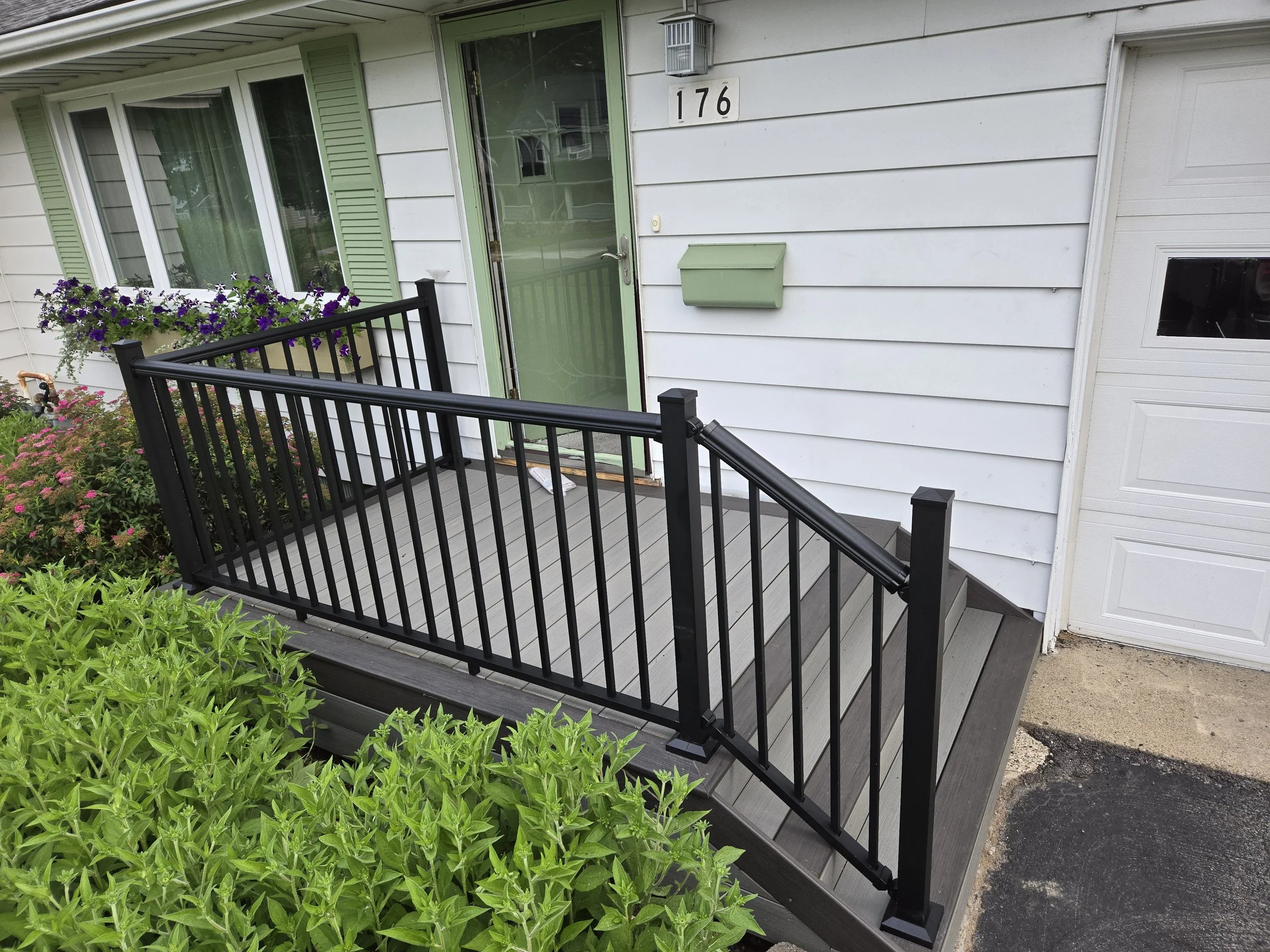 Front porch with gray wooden stairs and black metal railing, white house siding with a green door and window shutters, purple flower box, green mailbox, and a white garage door.