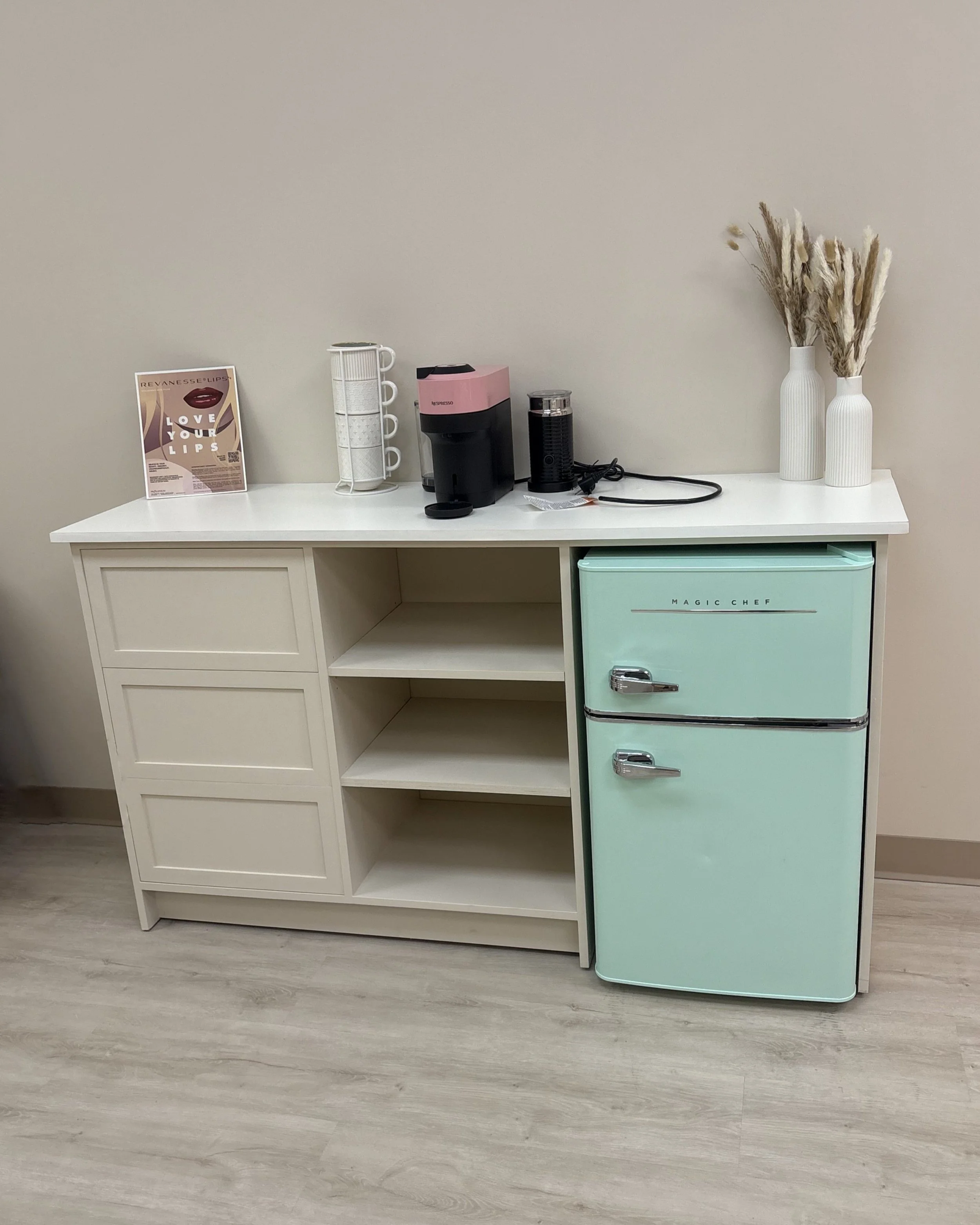 A white sideboard with open shelves and a retro mint green mini refrigerator. On top, there are two white vases with dried pampas grass, a coffee maker, a water filter, and a display stand with a flyer.