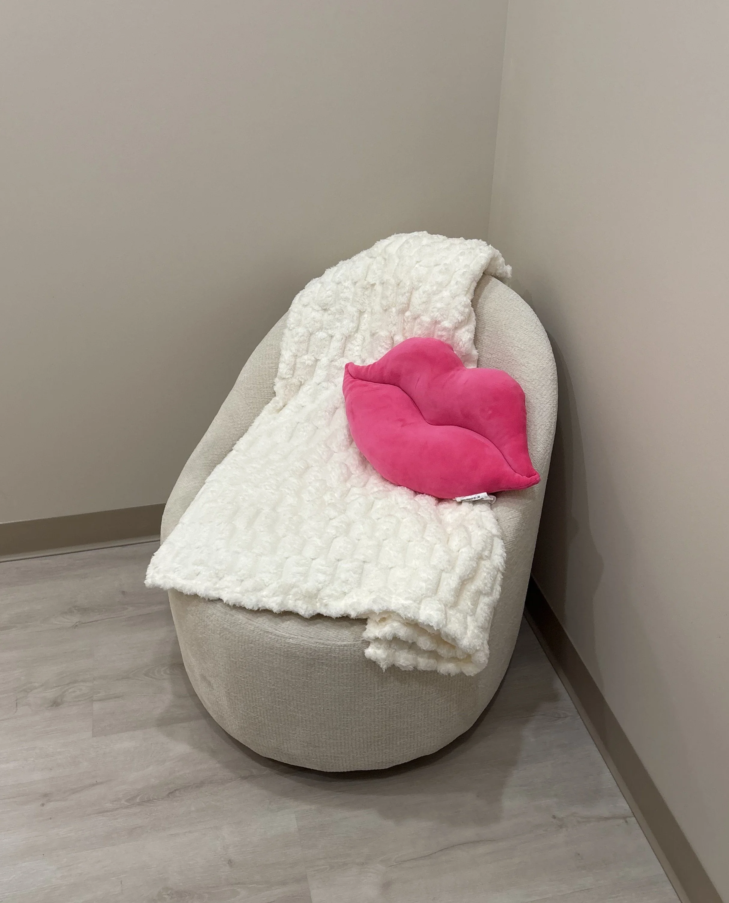 A beige upholstered chair with a soft white textured throw blanket and a pink lips-shaped cushion, placed in the corner of a room.