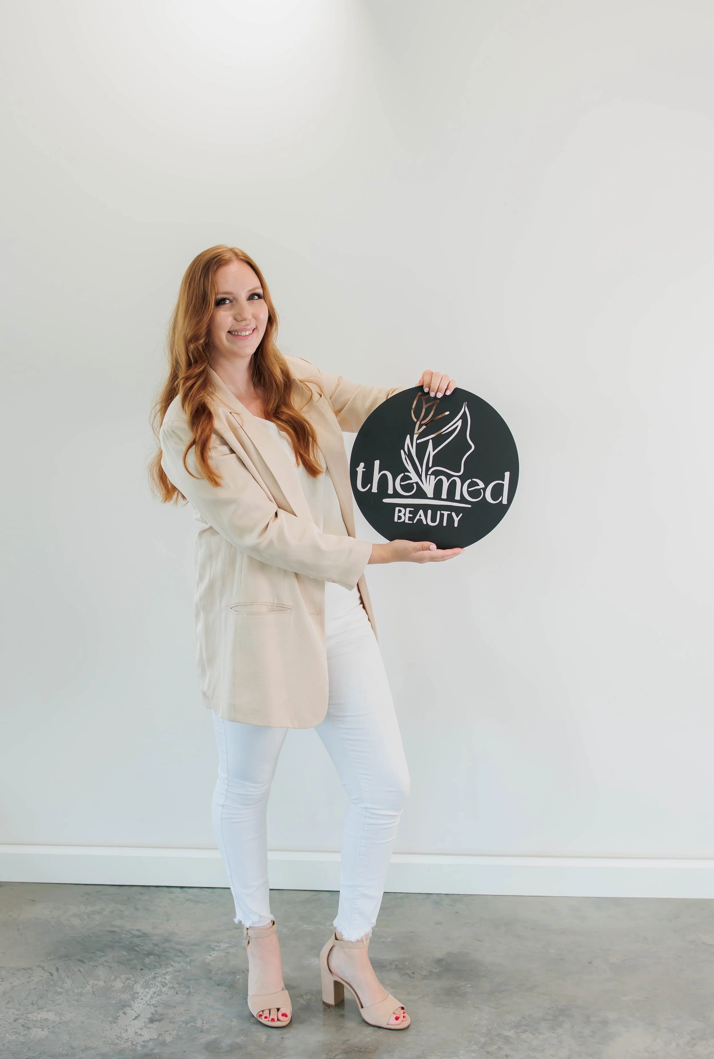 A woman with long red hair wearing a beige blazer, white pants, and beige high heels, smiling and holding a round black sign with the logo and text "the med BEAUTY" on a plain white background.