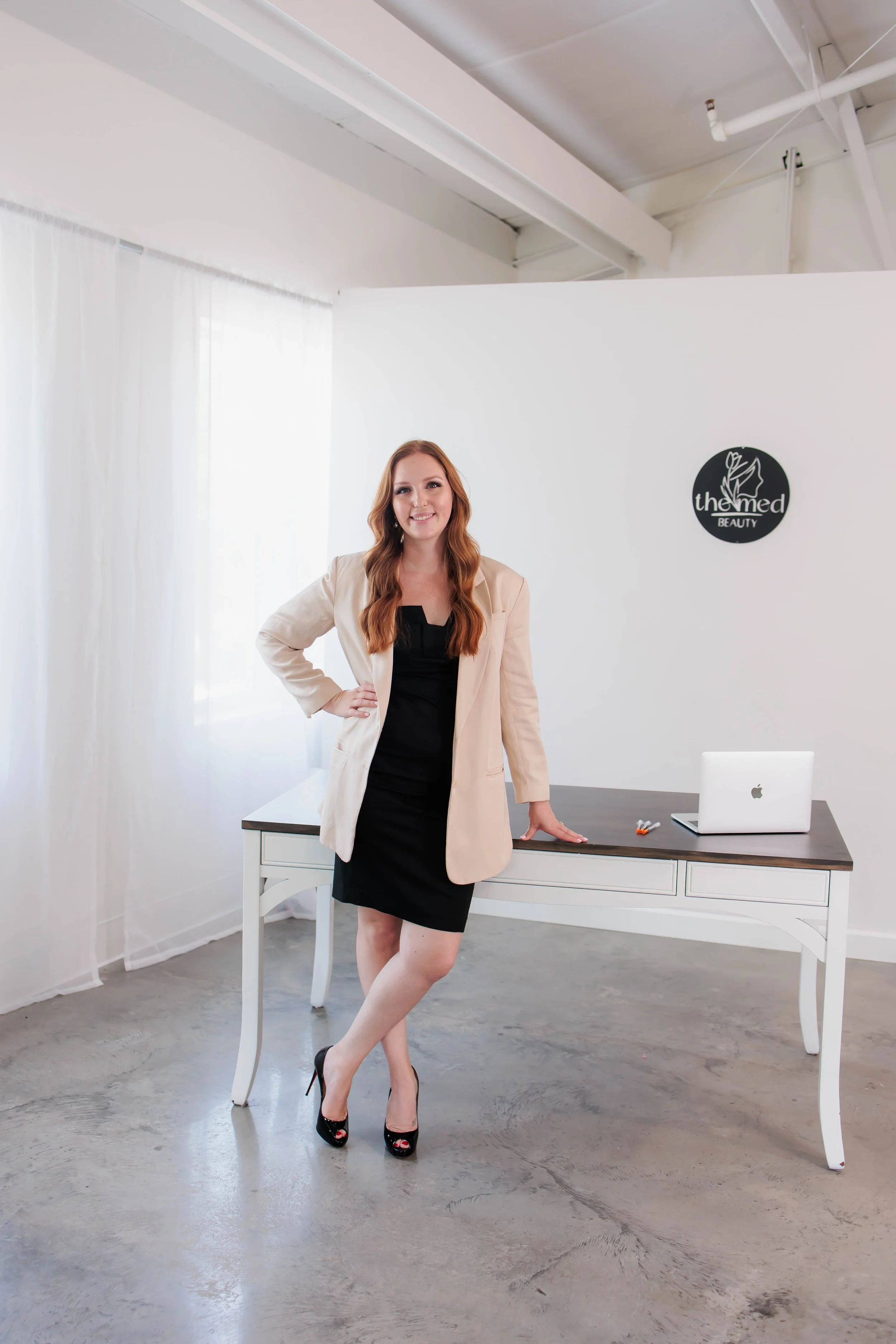 A woman with long red hair in a black dress and beige blazer standing in a modern, bright office space next to a white table with a laptop, in front of a white wall with a logo that reads 'The Med Beauty'.