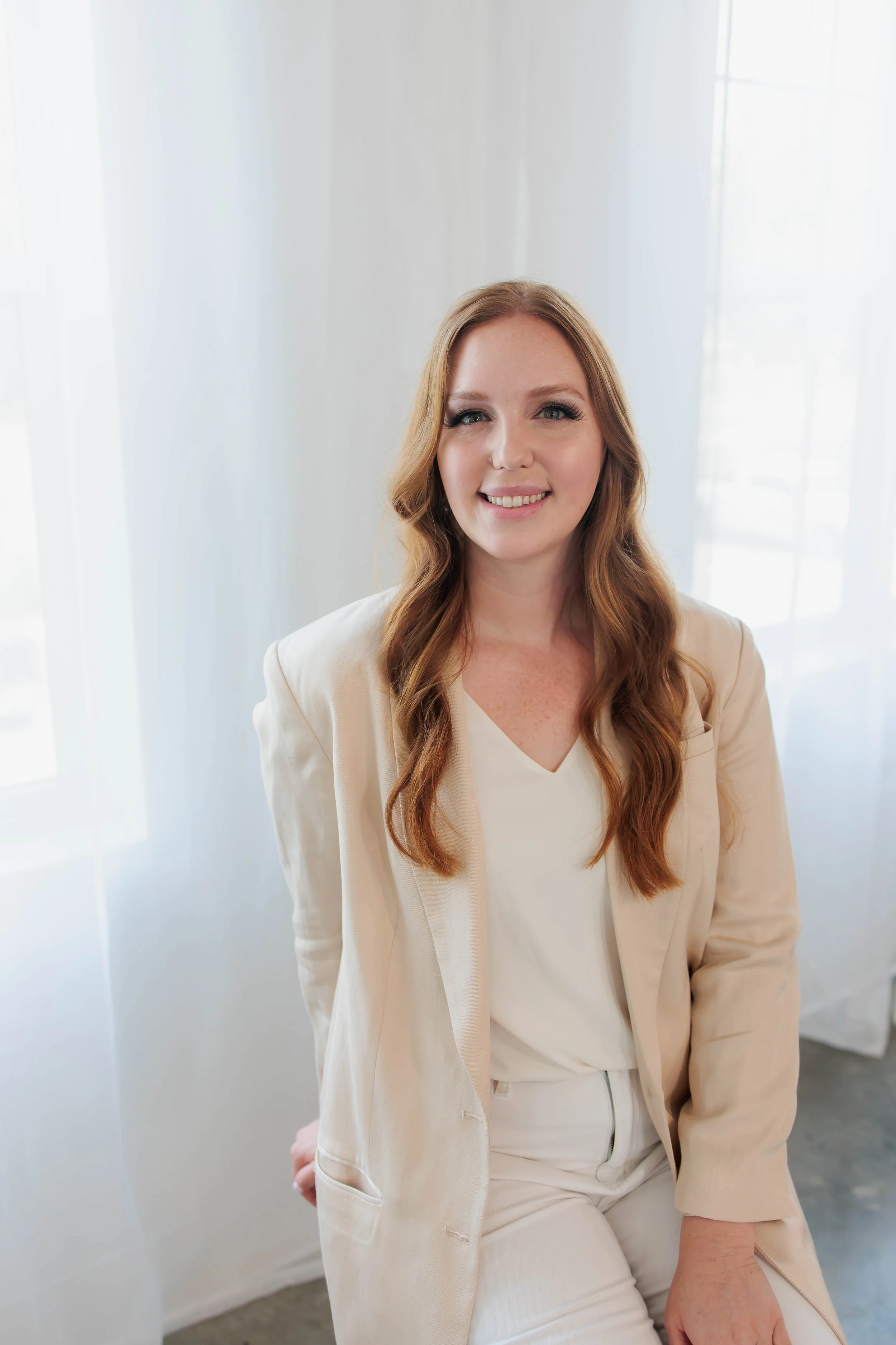 A woman with long red hair, dressed in white, smiling in front of a window with sheer white curtains.