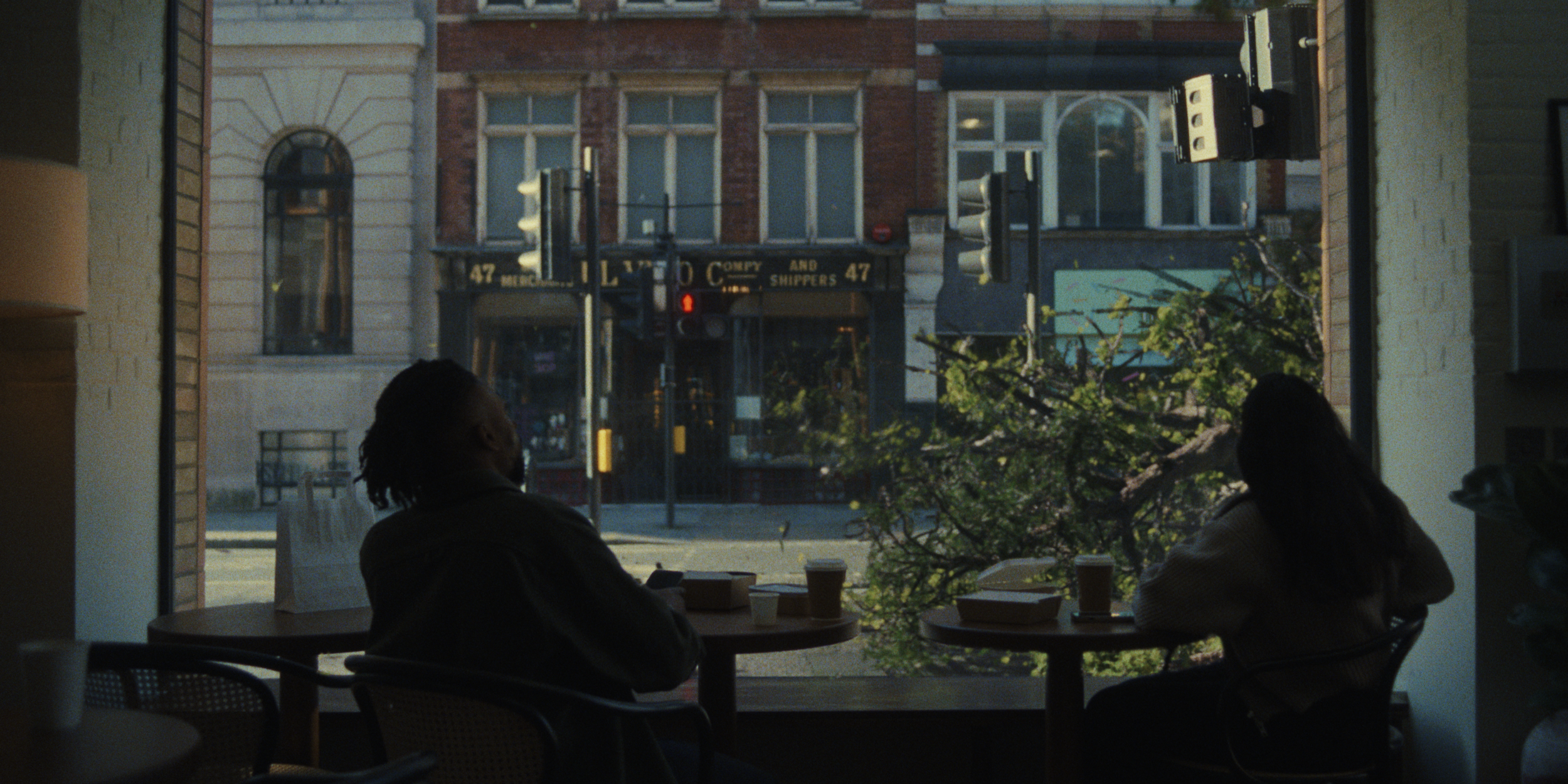 Storm Elfrink - Canopy - Pack4Good - Inside a coffee shop, with two people seated at a table near a large window. The view through the window shows a street scene with buildings and trees, and a pedestrian crossing signal.