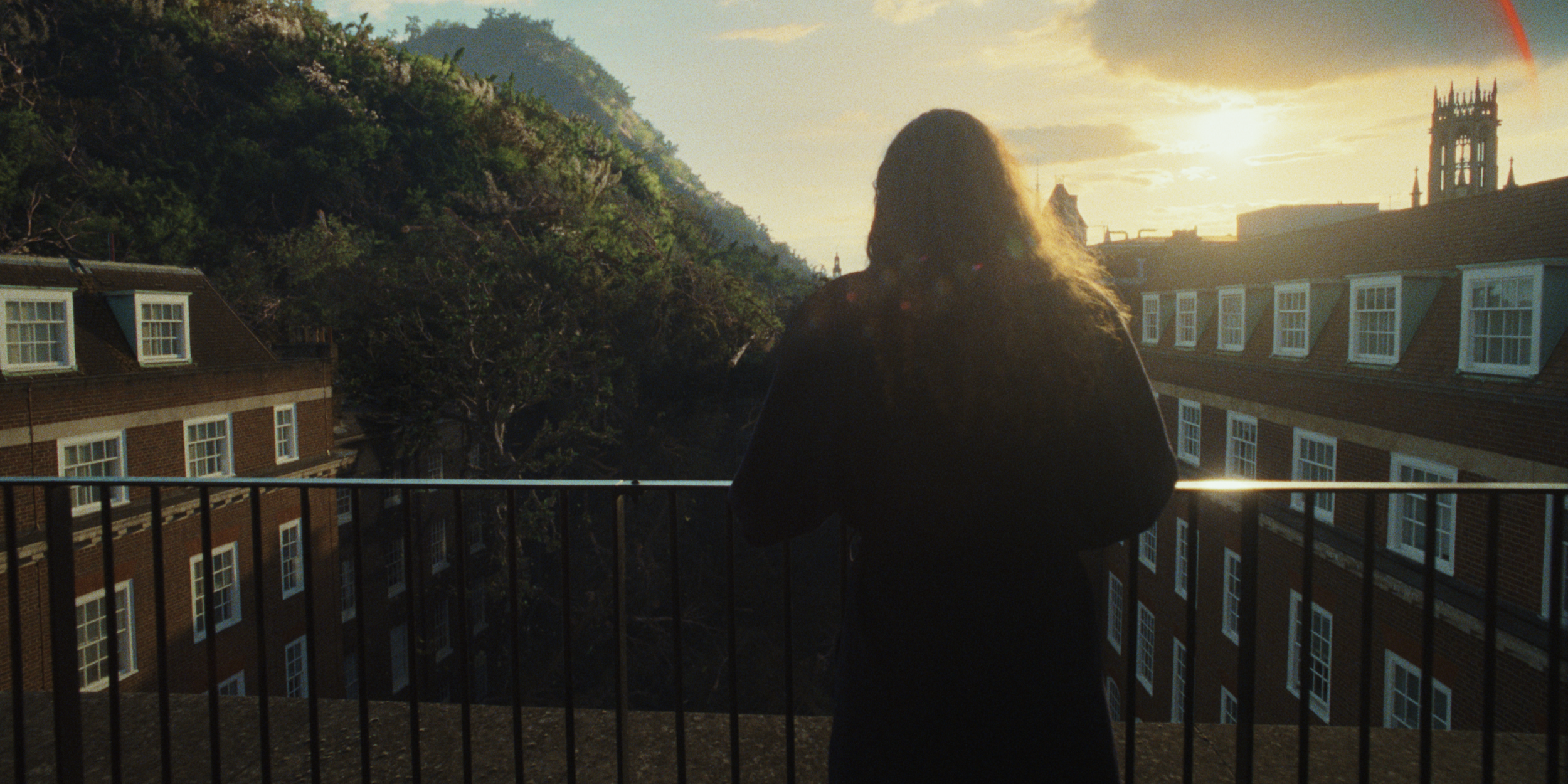 Storm Elfrink - Canopy - Pack4Good - A woman standing on a balcony overlooking buildings and trees during sunset, with the sun shining through clouds in the background.