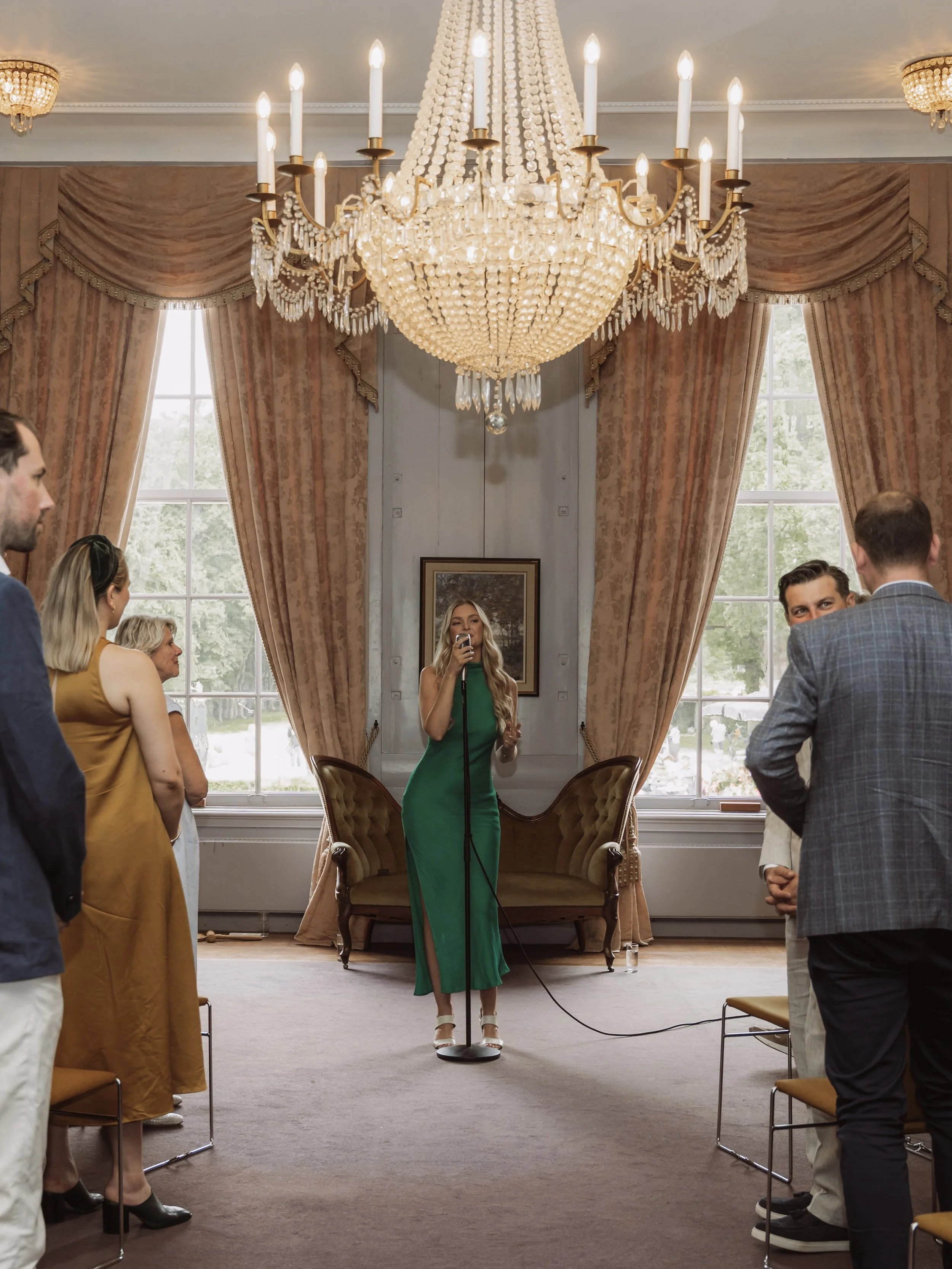 Een vrouw in een groene jurk zingt in een elegante kamer met een grote kristallen chandelier, omringd door enkele mensen die naar haar luisteren.