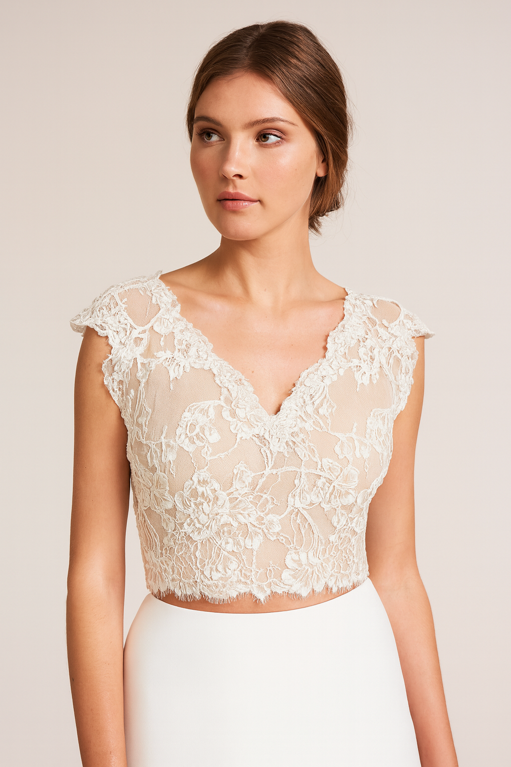 A young woman with short brown hair, wearing a cream lace crop top with floral embroidery and a high-waisted white skirt, standing against a plain beige background.