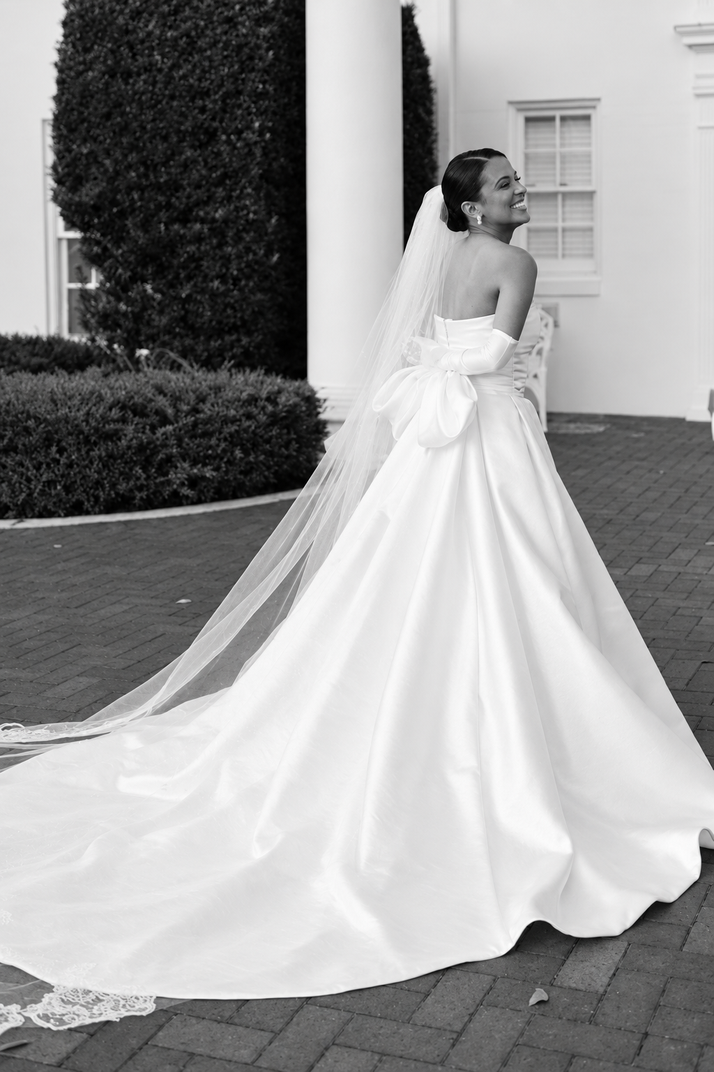 Sira D’ Pion bride in a classic satin ball gown, showing the full skirt, bridal bow, and sweeping train in a black-and-white wedding portrait.