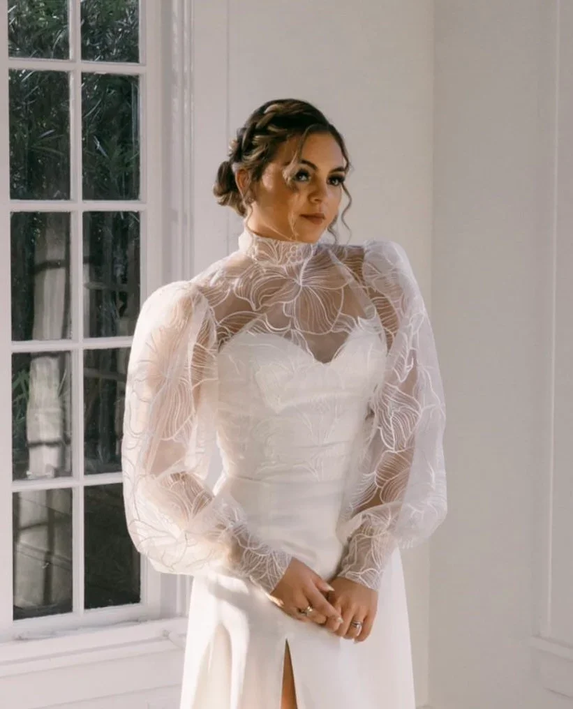 A woman in a white wedding dress with puffed sleeves and a lace embroidered sheer top, standing indoors near a window with trees visible outside.
