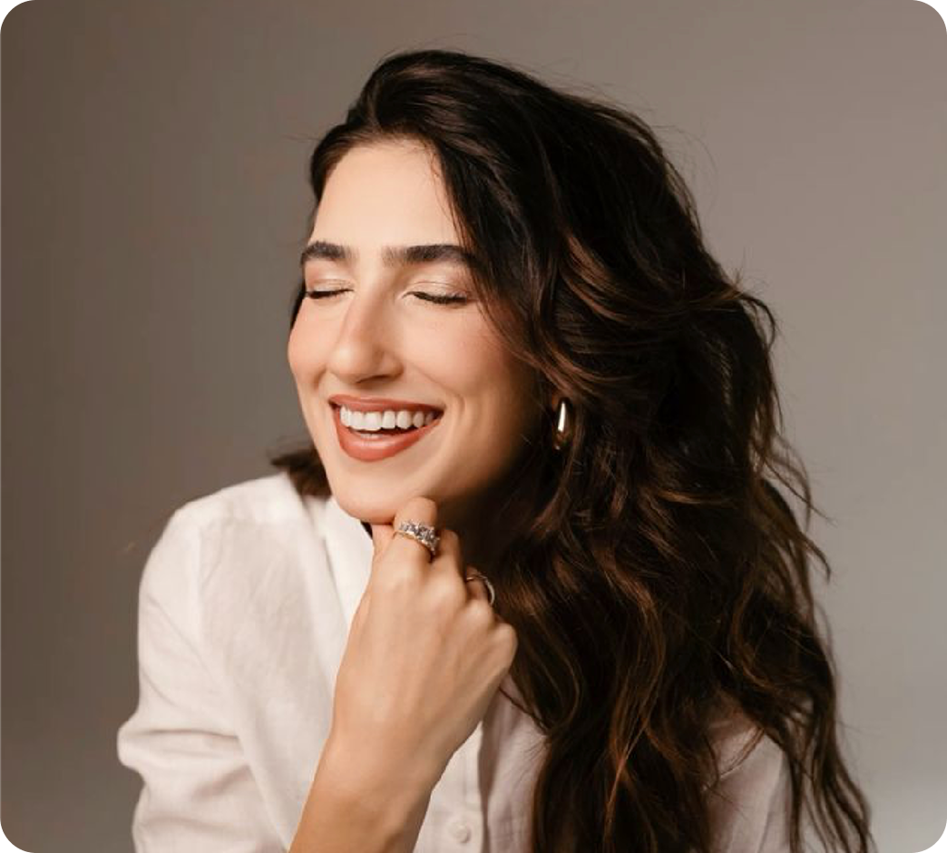 Mujer sonriendo con ojos cerrados, cabello castaño oscuro y rizado, usando pendientes grandes y anillos, con blusa blanca.