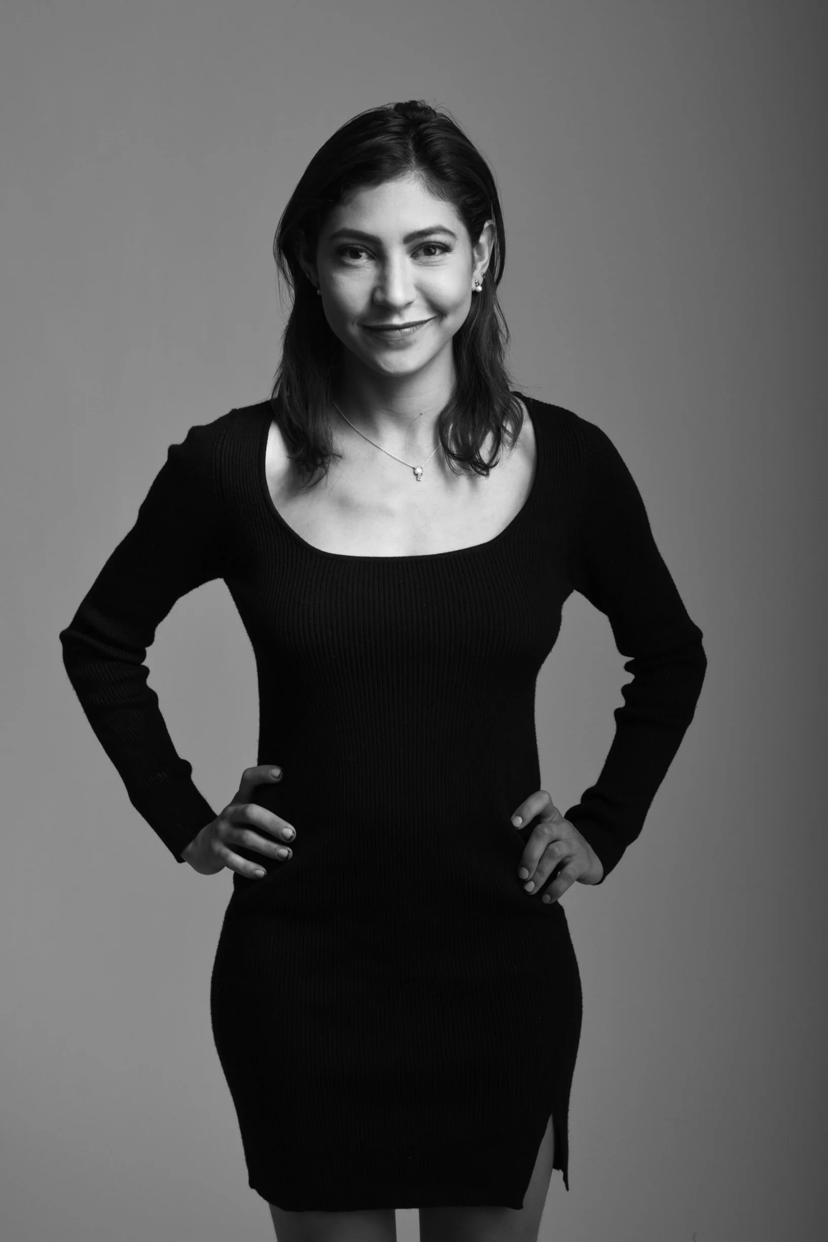 Retrato en blanco y negro de una mujer con cabello castaño oscuro, usando un vestido ajustado y un collar, con las manos en las caderas, sonriendo suavemente.