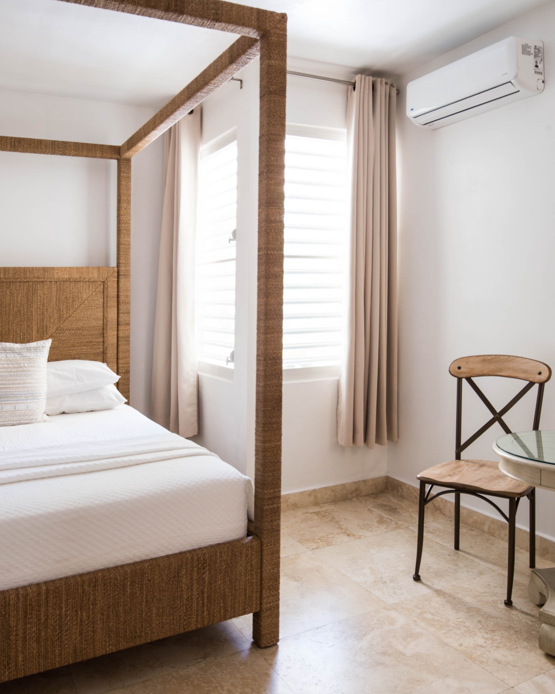 Minimalist bedroom with a wicker canopy bed, white bedding, air conditioning unit, small chair, and round table near windows with curtains.
