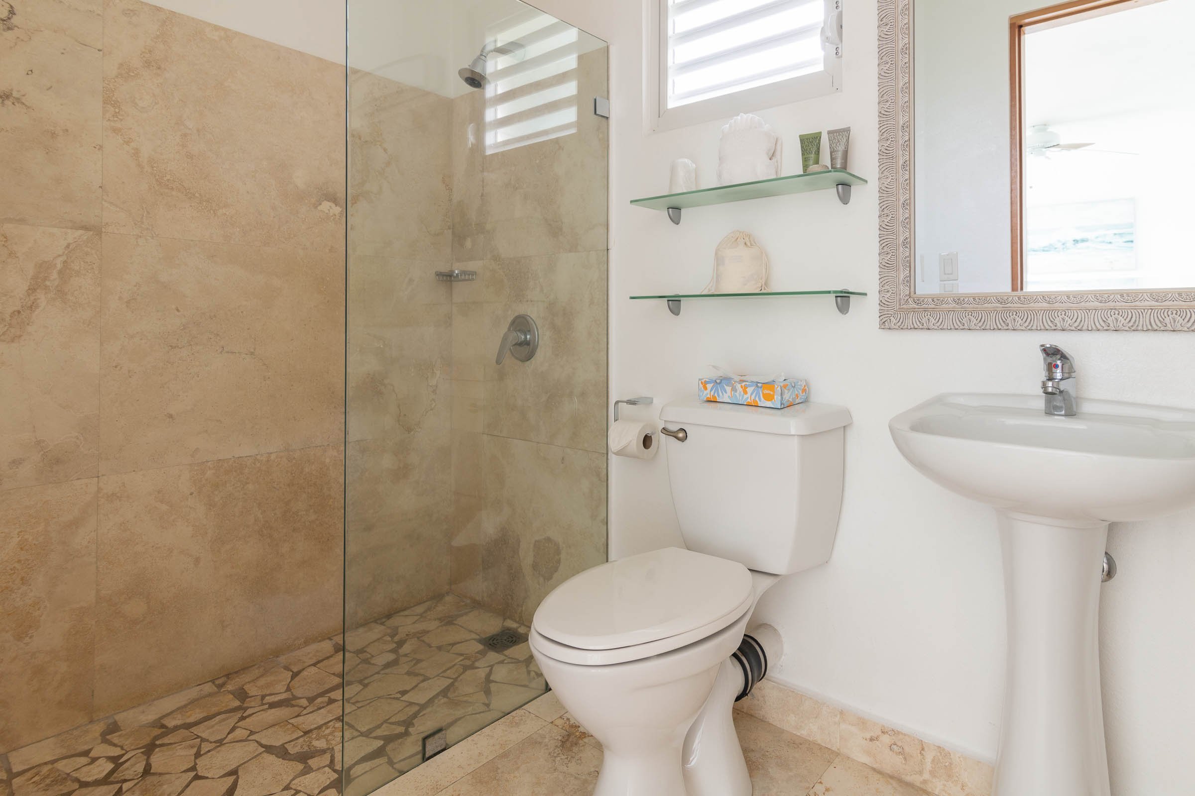 Modern bathroom with a glass-enclosed shower, toilet, pedestal sink, and glass shelves with toiletries.