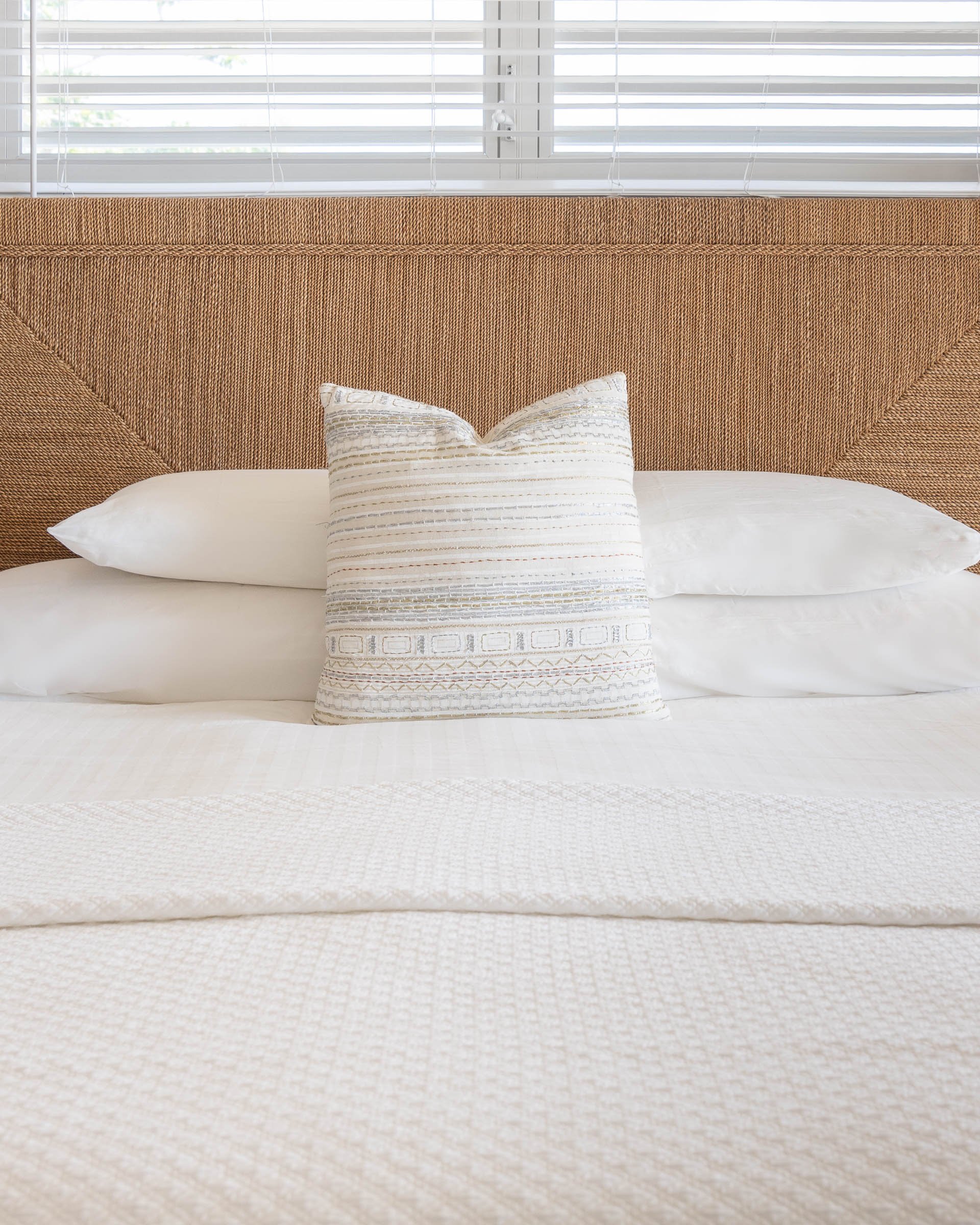 Bed with white bedding, patterned pillow, and wicker headboard, under a window with blinds.