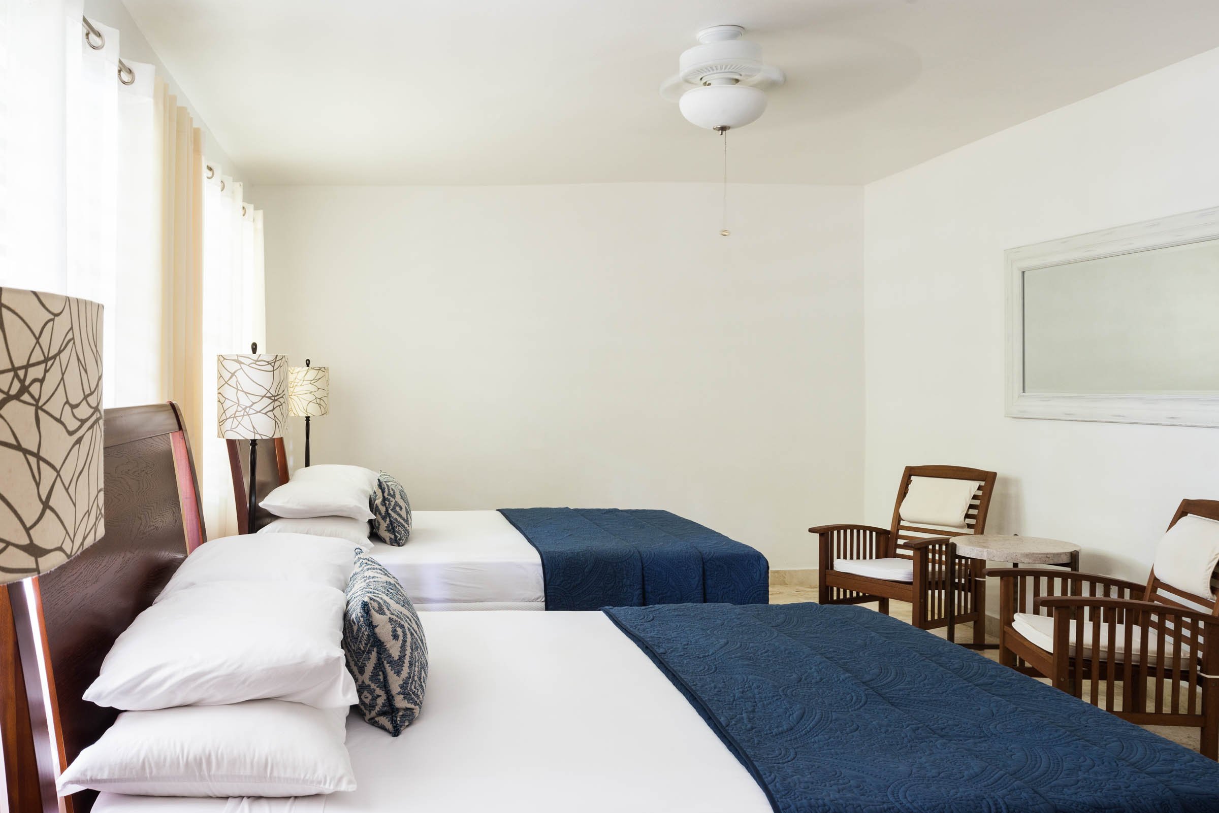 Hotel room with two double beds, blue bed runners, wooden headboards, two modern lamps, a ceiling fan, two wooden chairs, a small round table, and a wall mirror.
