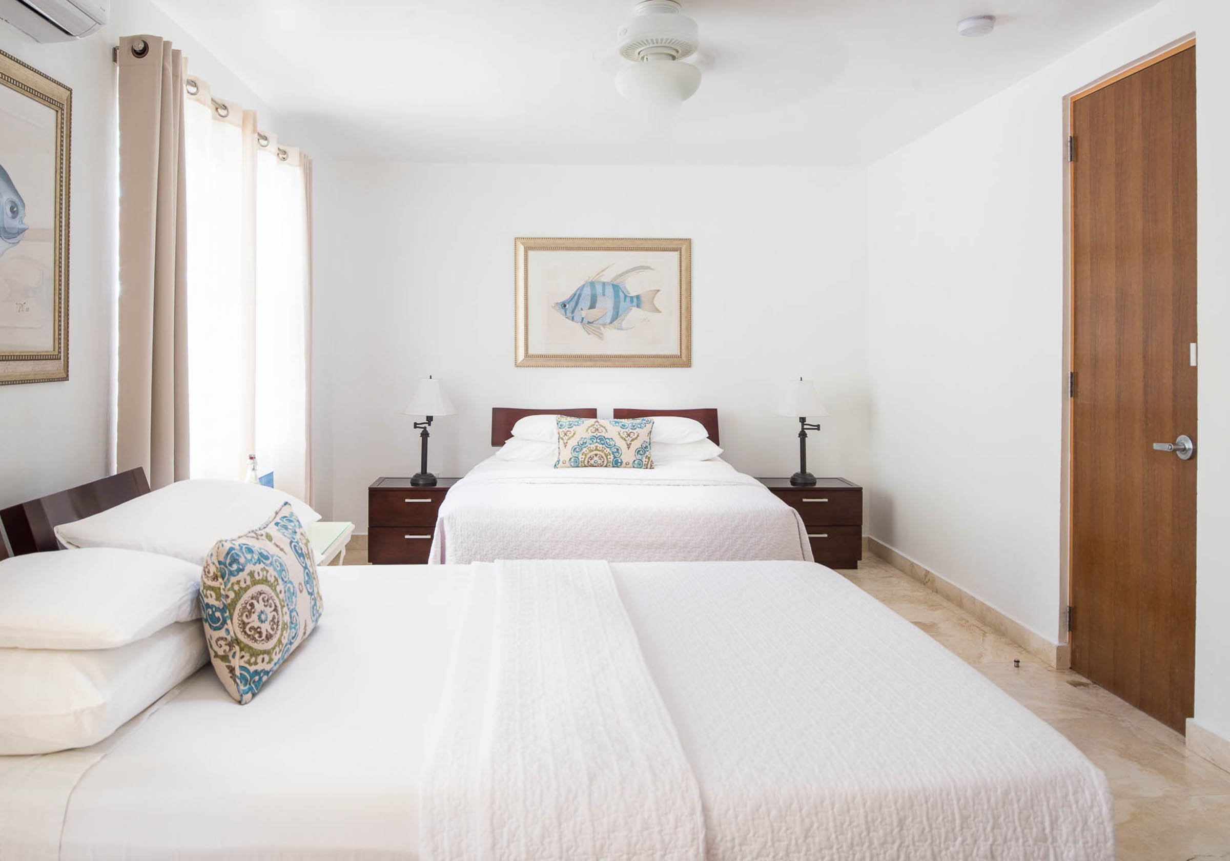 Hotel room with two queen beds, white bedding, decorative pillows, framed fish art, bedside tables, lamps, and wooden door.