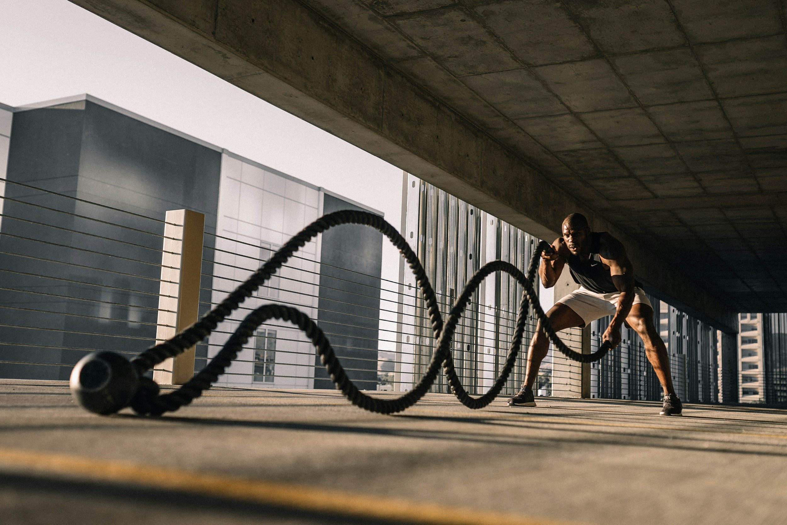 A man performing a battle rope workout under a bridge in an urban area, with city buildings in the background.