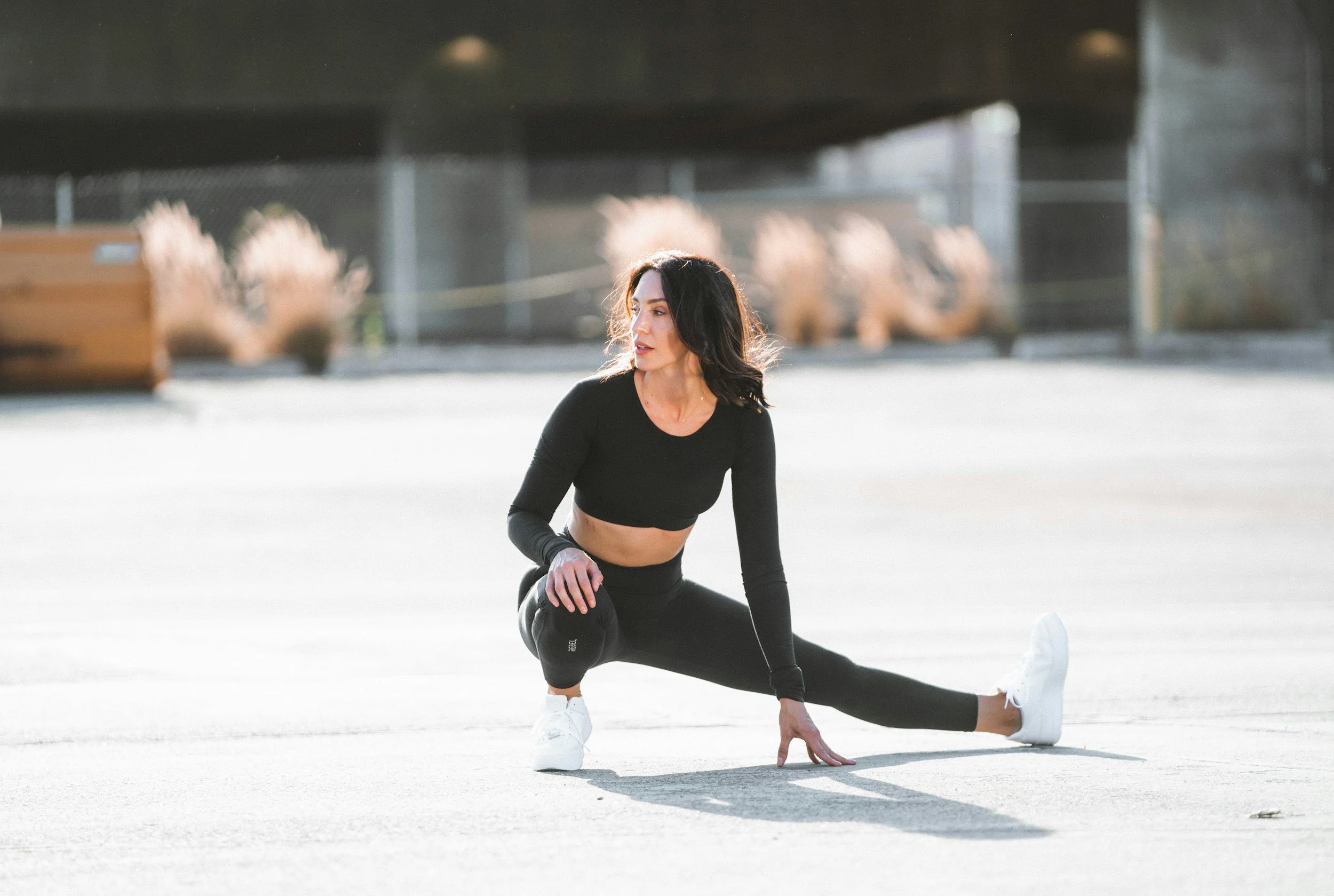 A woman in black athletic clothes and white sneakers stretching outdoors on a concrete surface, with plants and an overpass in the background.