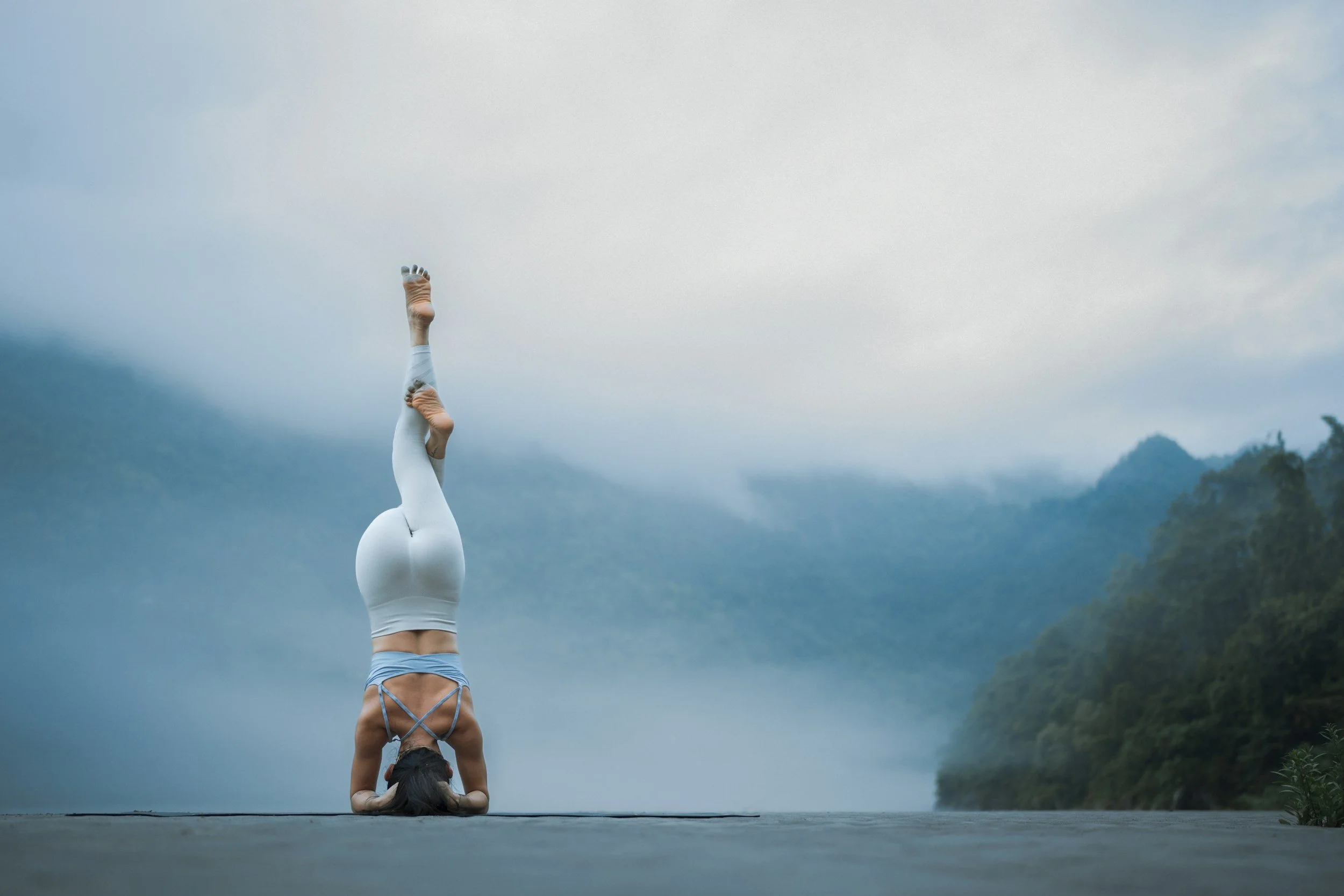 A woman practicing yoga in a handstand on an outdoor platform with foggy mountains and trees in the background.